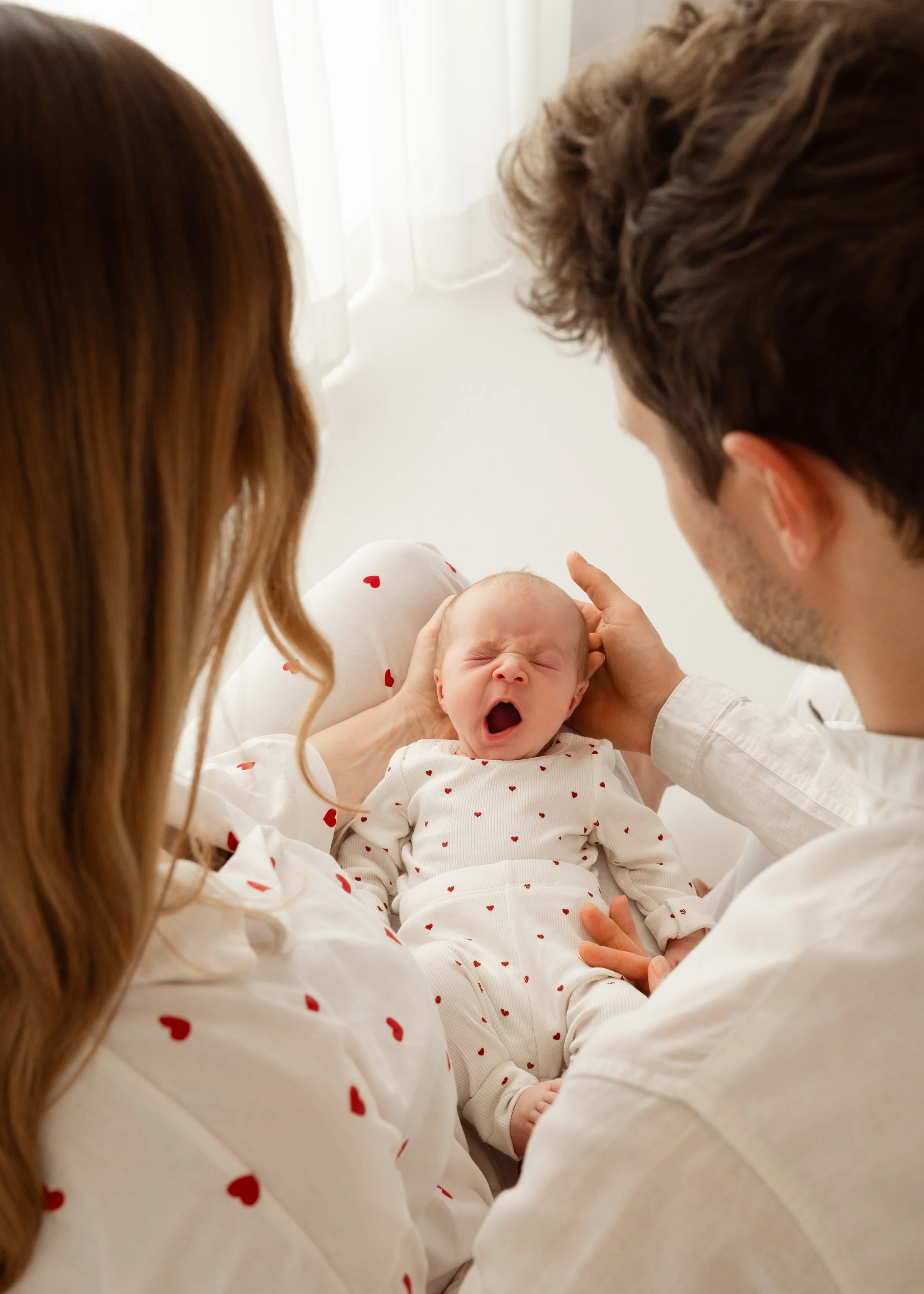 Ein kleines Baby, das von einer Frau und einem Mann anscheinend die Eltern, gehalten wird. Das Baby trägt ein weißes Kleid mit roten Herzchen, während die Eltern ebenfalls Kleidungsstücke mit roten Herzen tragen.