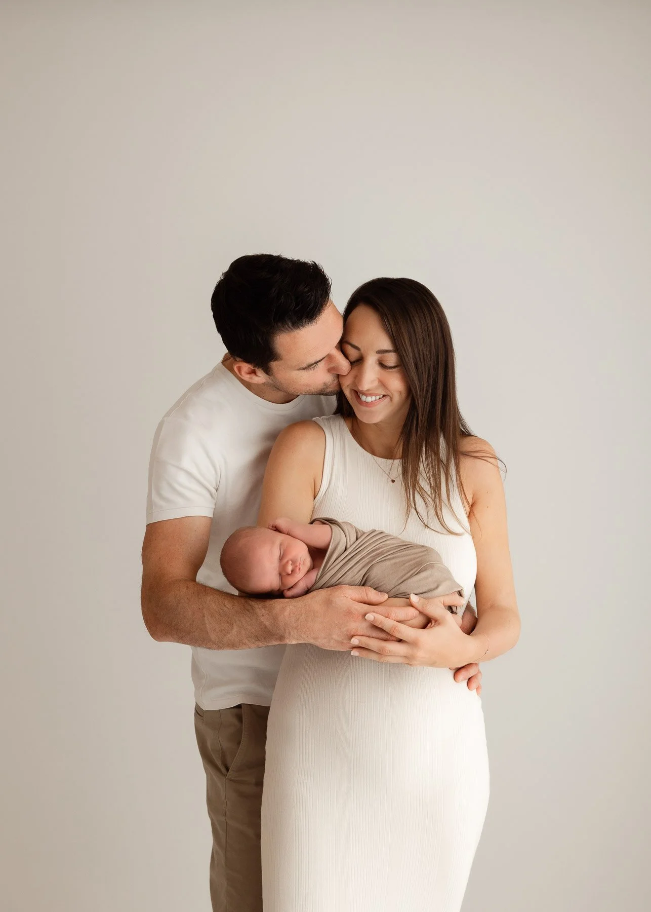 Ein glückliches Paar hält ein schlafendes Baby, während der Mann die Frau küsst. Die Szene ist vor einem neutralen Hintergrund. Es wirkt wie eine Familienaufnahme nach der Geburt.