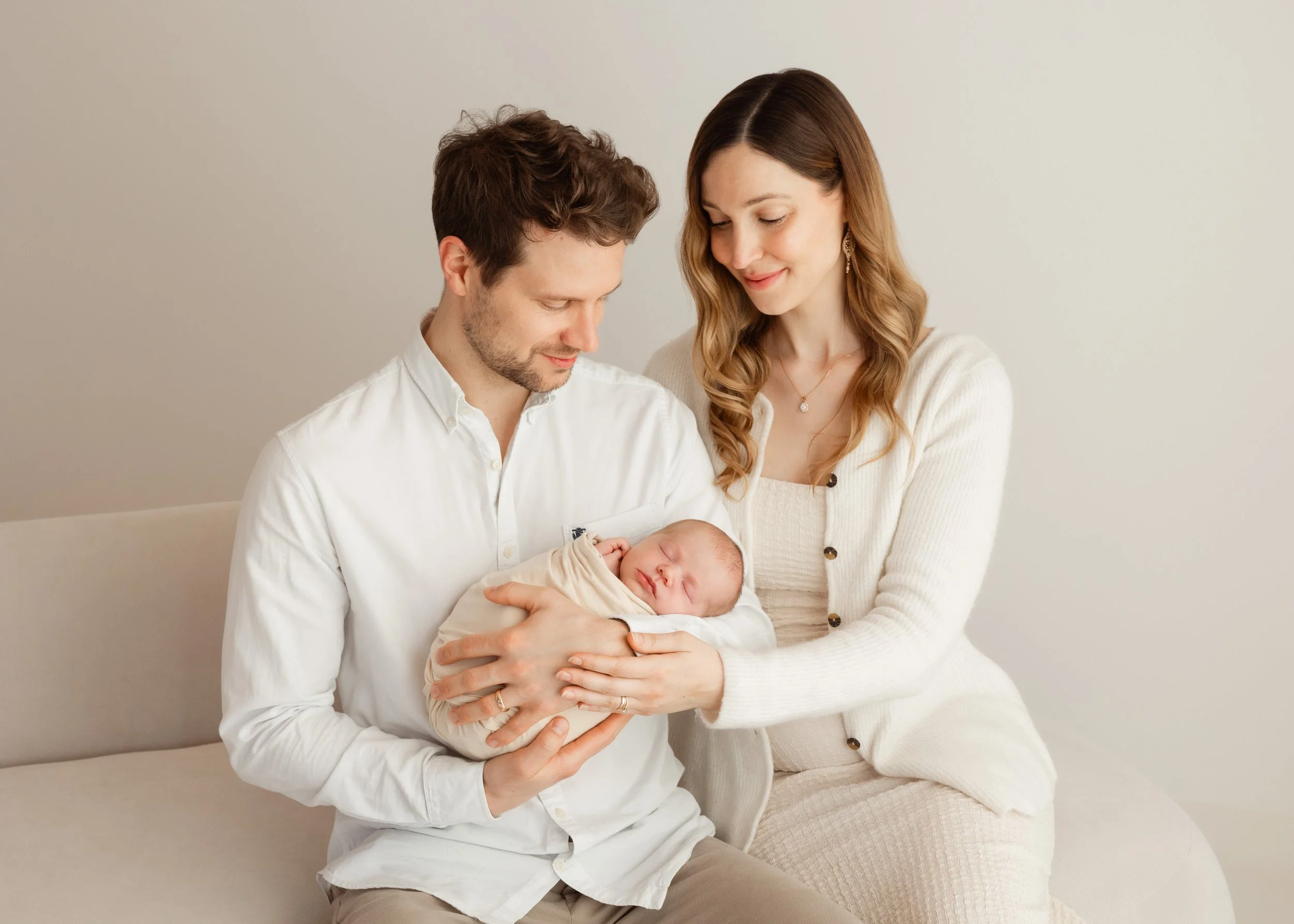 Ein glückliches Paar hält ein schlafendes Baby in den Armen, während sie auf einer Couch sitzen, in einem hellen, minimalistischen Raum.