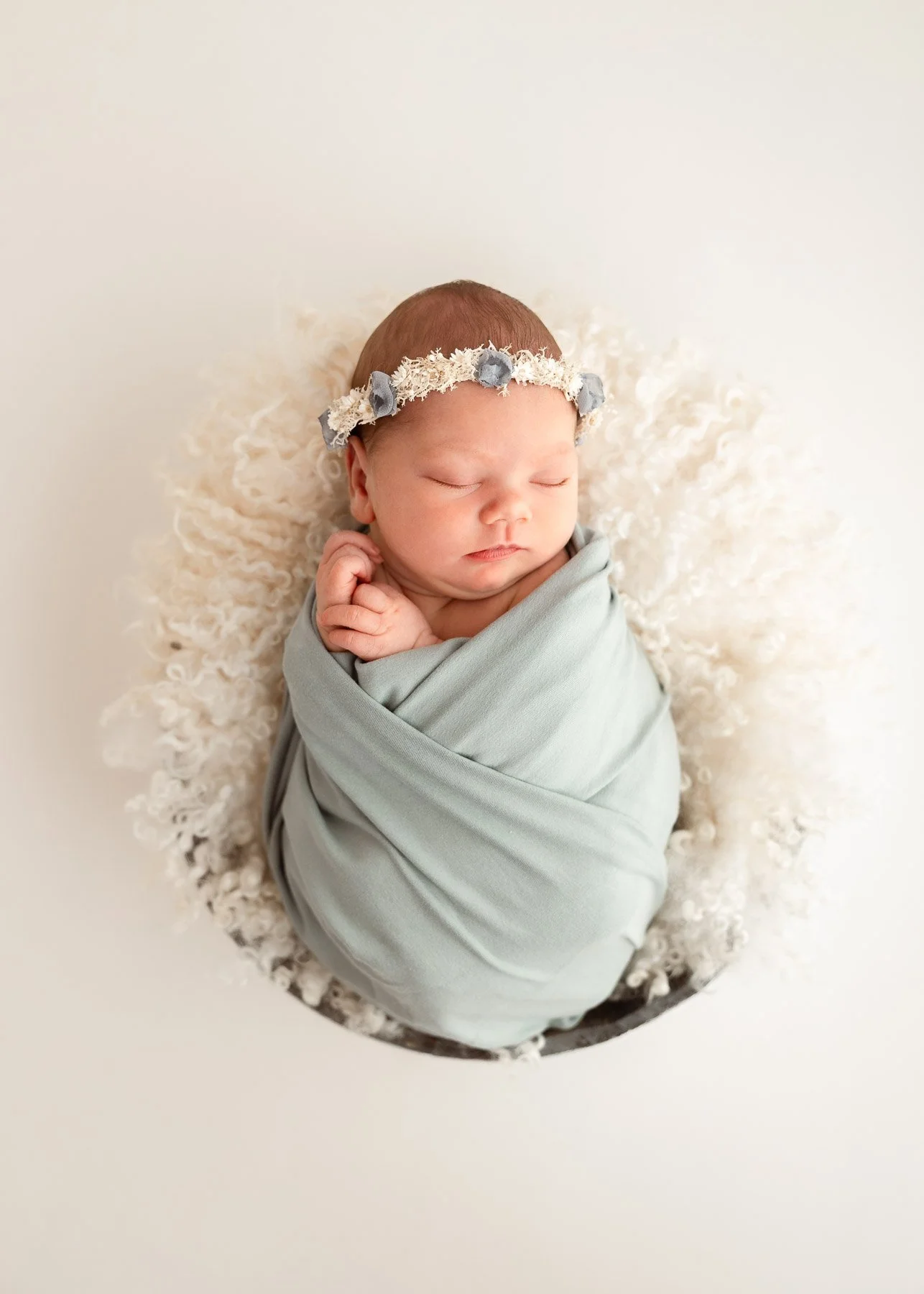 Ein schlafendes Neugeborenes, eingewickelt in einen hellgrauen Stoff, mit einem weißen und blauen Blumen-Haarband, auf einer weißen, fluffy Unterlage.