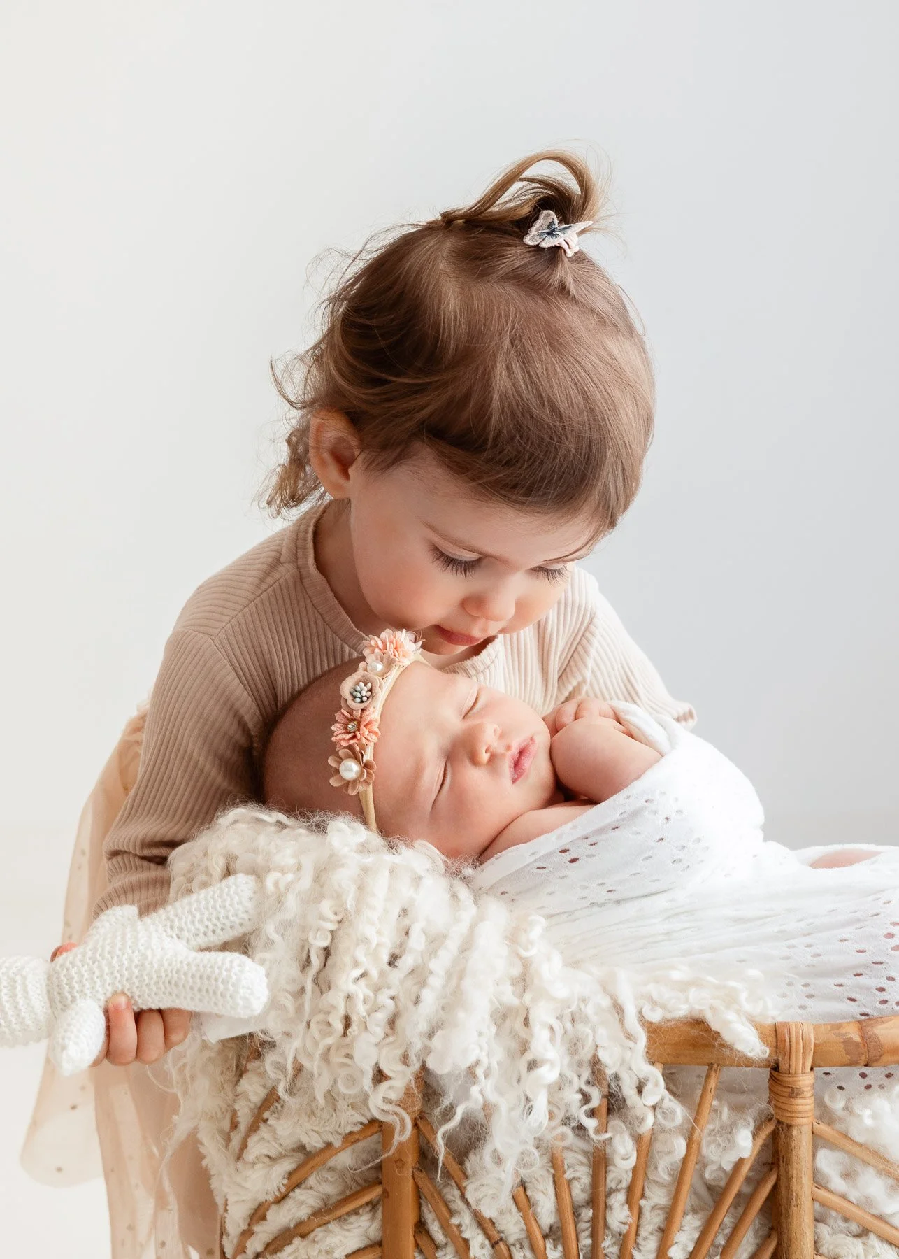 Ein kleines Mädchen hält ein schlafendes Baby in einem Korb, beide sind liebevoll zueinander.