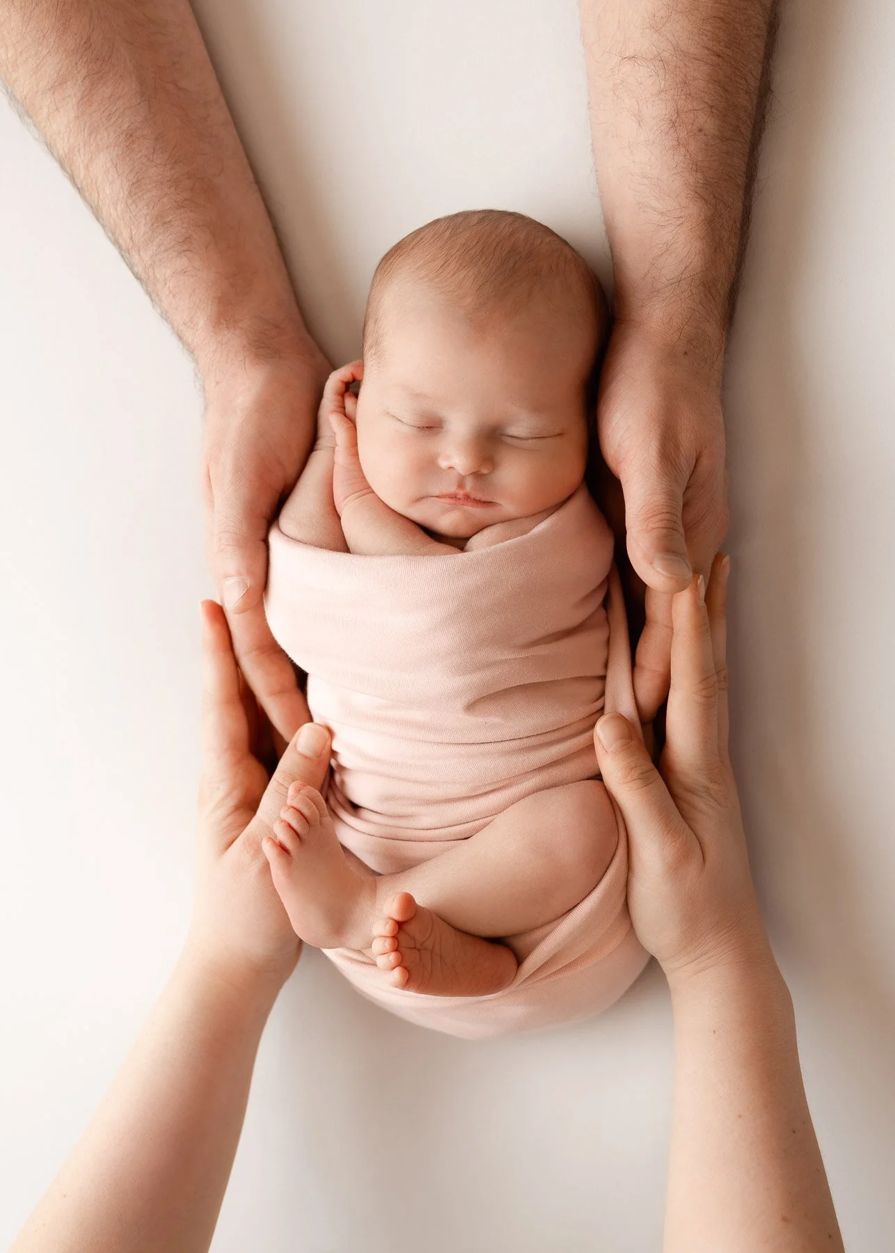 Ein schlafender Neugeborener in einen rosa Tuch eingewickelt, gehalten von verschiedenen Händen, auf weißem Hintergrund.