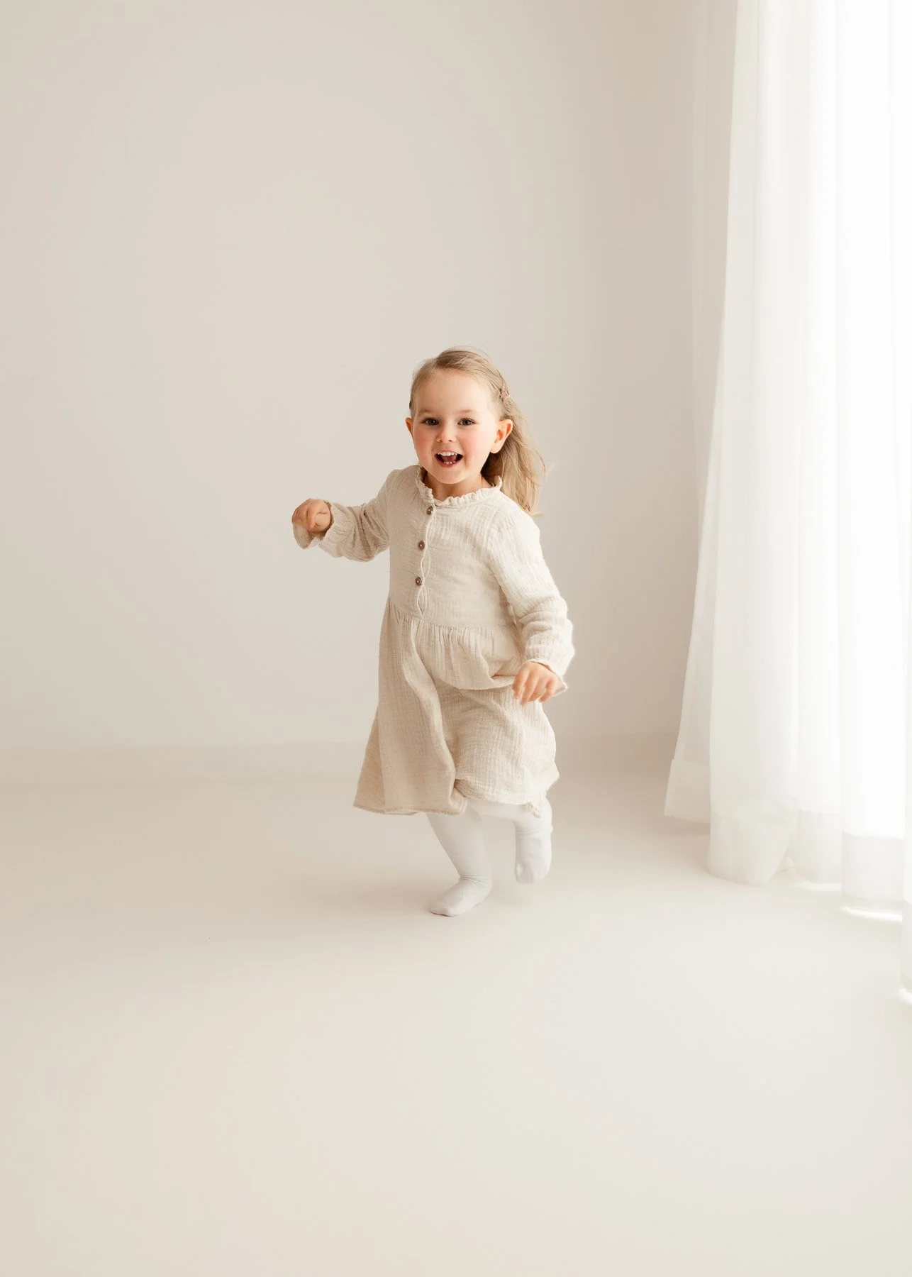 Kleines Mädchen läuft fröhlich in einem weißen Raum mit viel natürlichem Licht, trägt ein beige Kleid und weiße Socken.