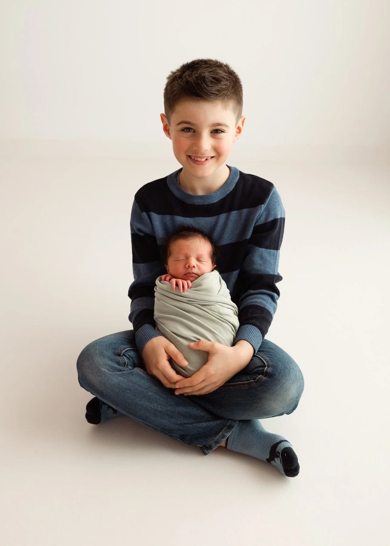 Ein lächelnder Junge hält ein in eine Decke gewickeltes Baby auf seinem Schoß, während er auf dem Boden sitzt.