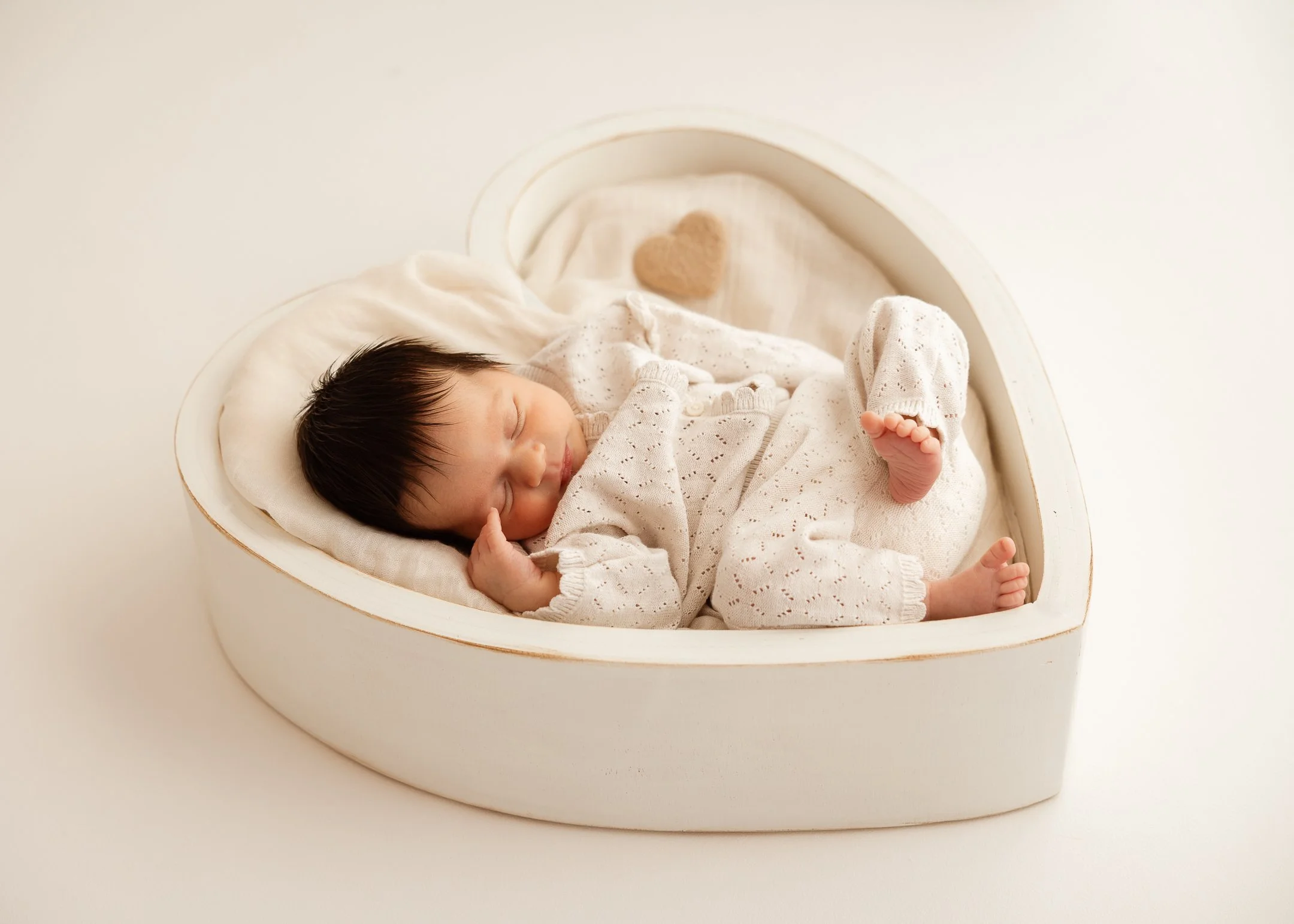 Ein schlafendes Baby liegt in einer herzförmigen weißen Kiste auf einem weichen Kissen, mit einem kleinen Plüschherz in der Kiste.