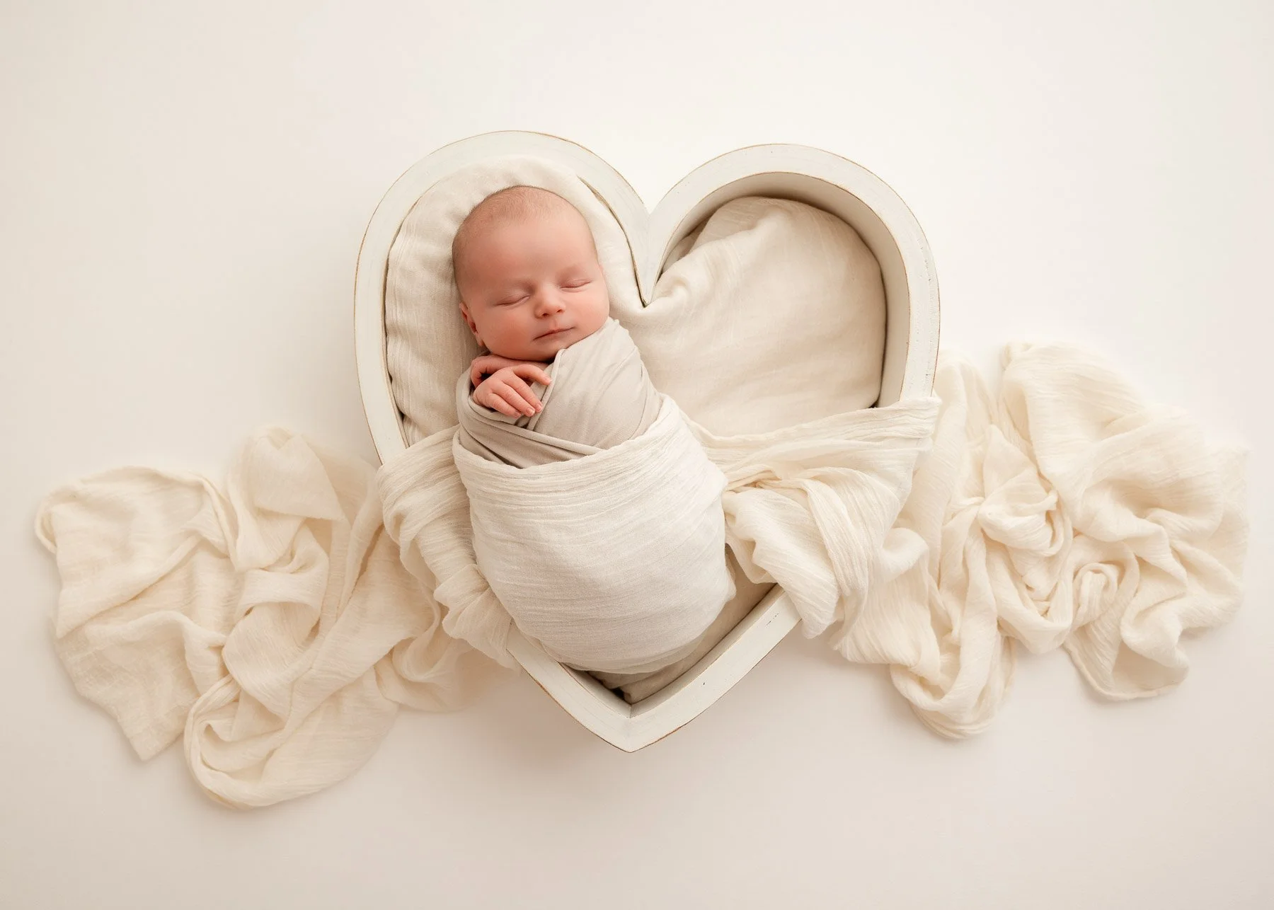 Ein schlafender Baby in einer weißen Herz-Schachtel, umgeben von cremefarbenen Tüchern.