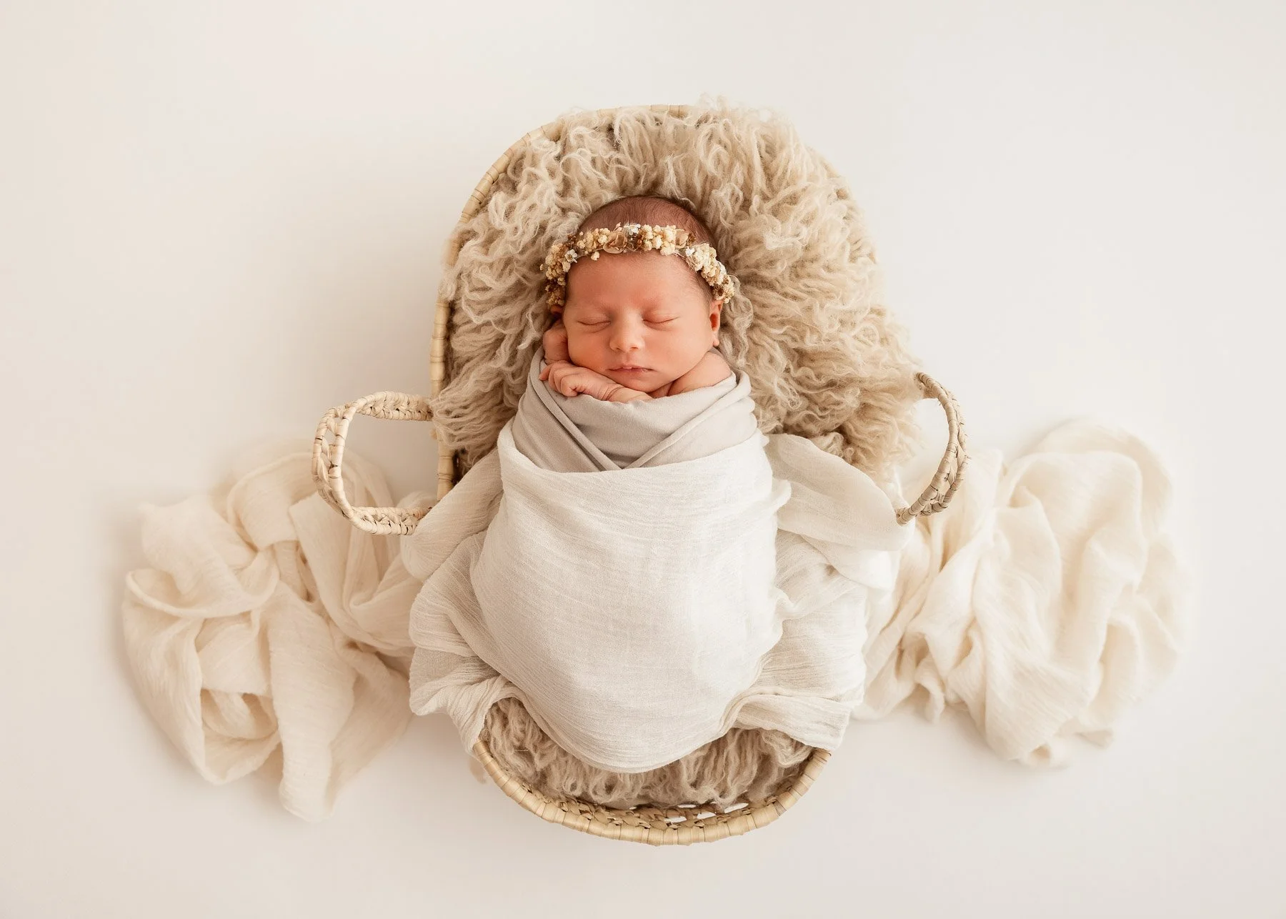 Ein schlafendes Neugeborenes in einem Körbchen, eingewickelt in weiße Tücher, mit einem Blumenkranz am Kopf, auf einem weichen Teppich.
