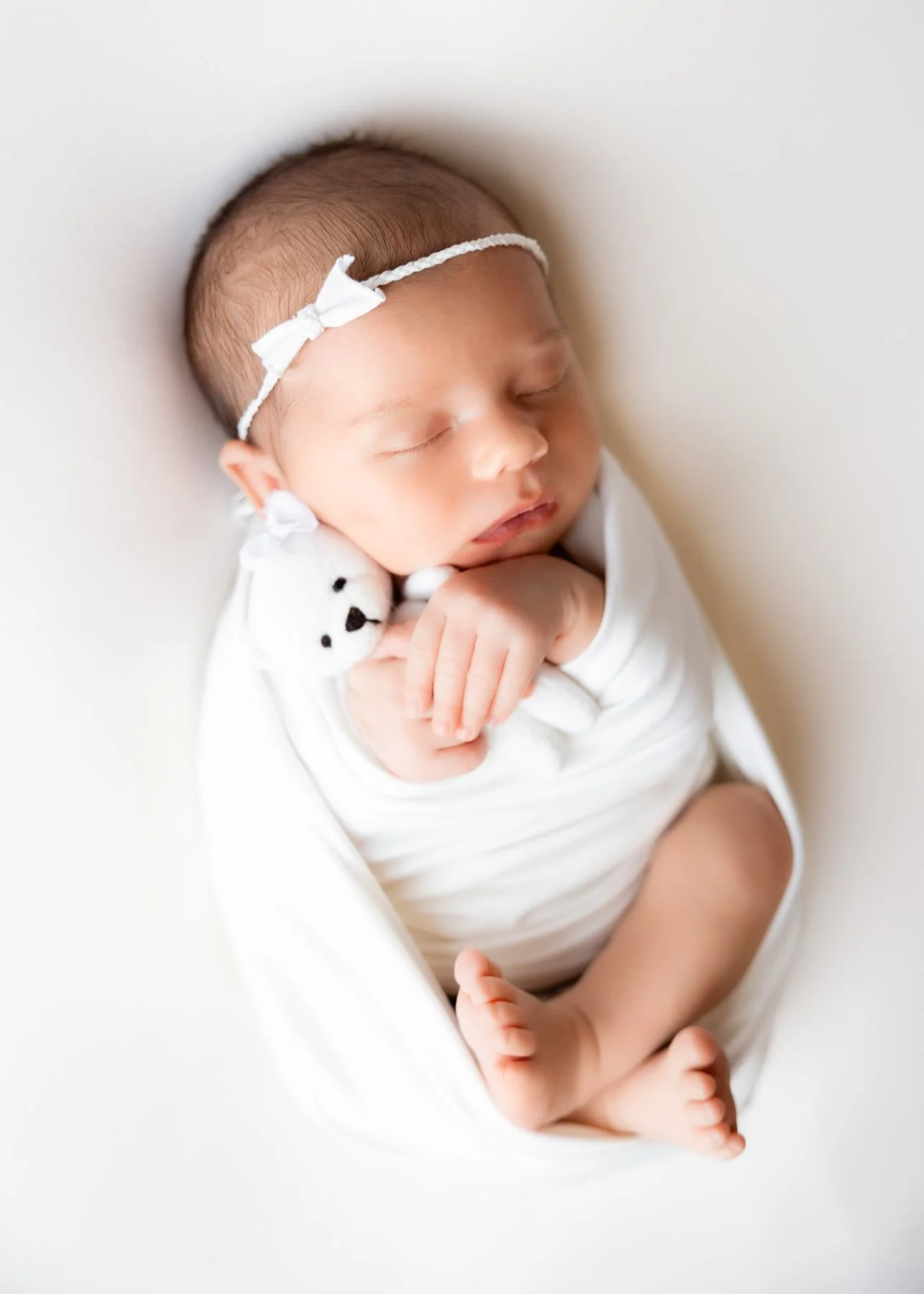 Schlafendes neugeborenes Baby mit weißen Kleid und Haarschmuck, hält einen kleinen weißen Teddybären auf der Schulter, auf weißem Hintergrund.