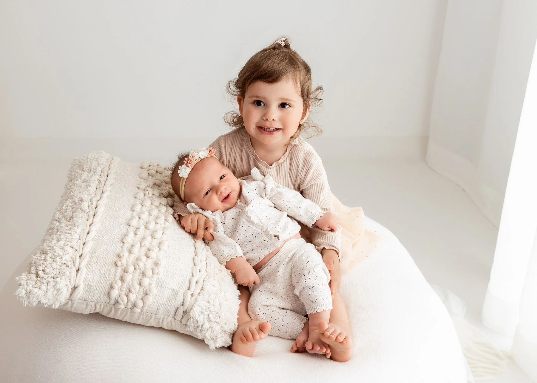 Ein kleines Mädchen hält ein Baby auf einem Bett mit weißen Bettwäsche und Kissen, beide tragen helle, schöne Kleidung. Das Mädchen lächelt freundlich.