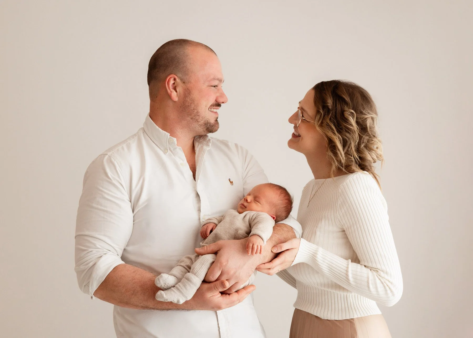 Ein Paar hält einen schlafenden Säugling und schaut sich glücklich an, vor einem neutralen Hintergrund.