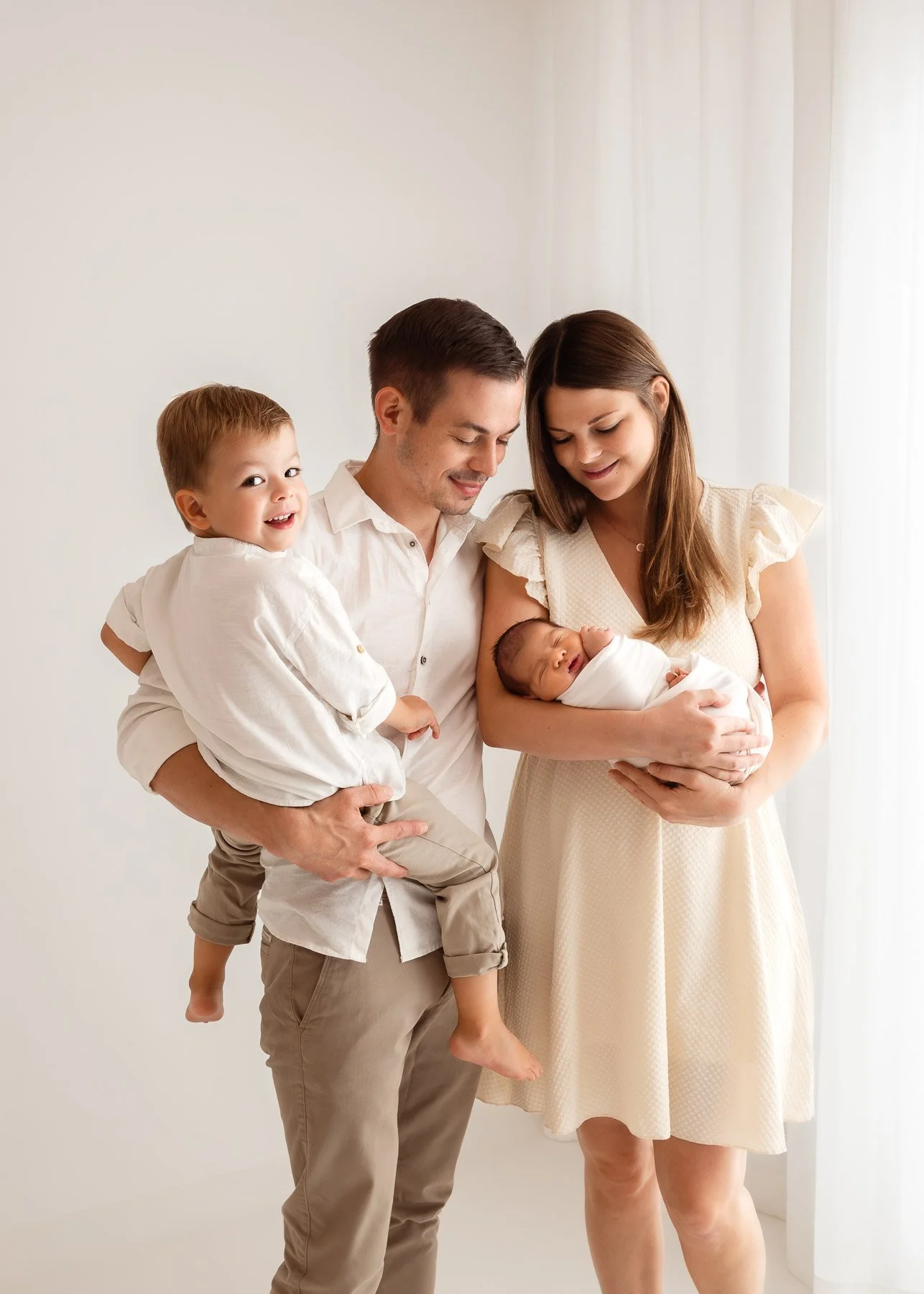 Familie mit Mutter, Vater, kleinem Kind, und neugeborenem Baby vor hellem Hintergrund.