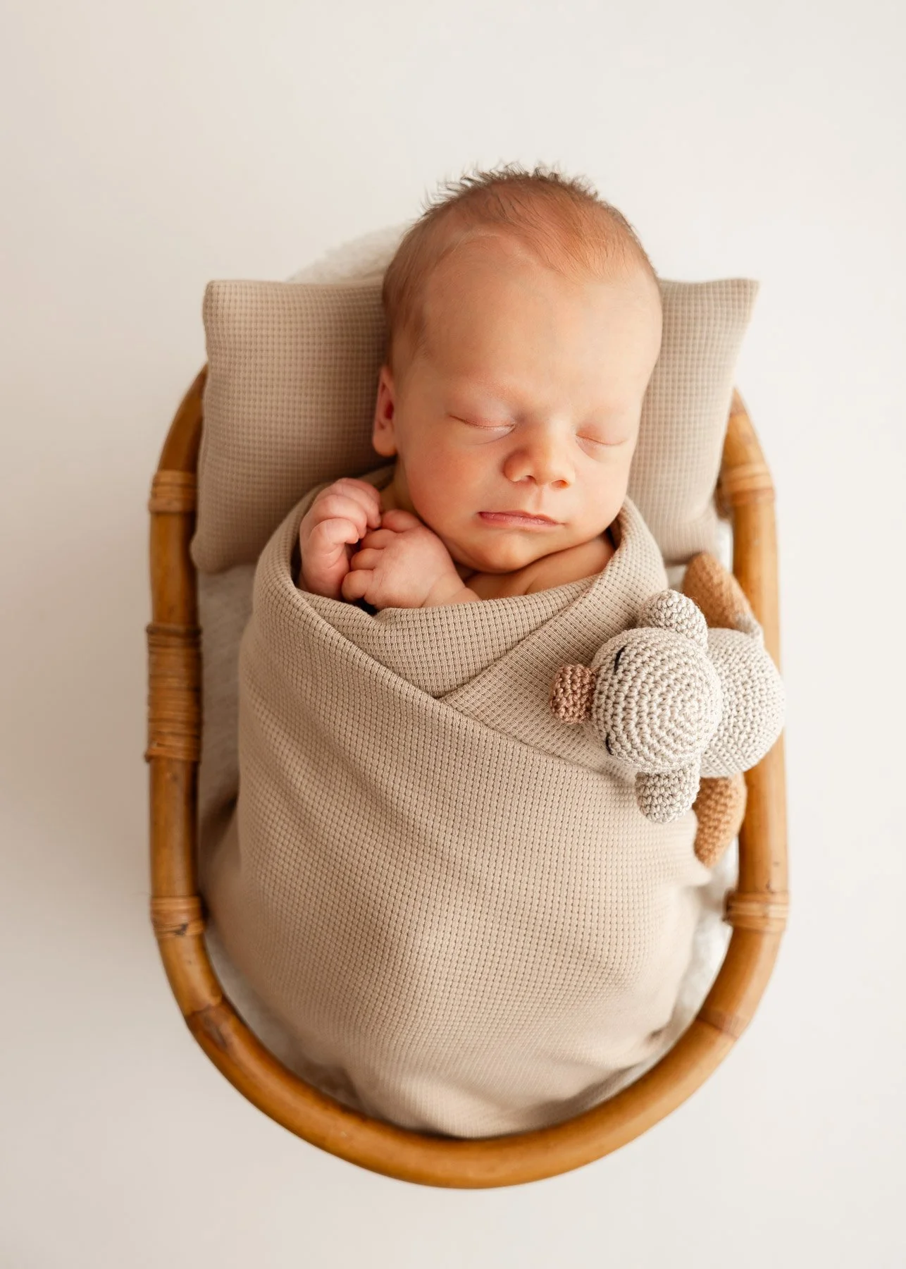 Ein schlafendes Baby in einem Korb, eingewickelt in eine beige Decke, mit einem kleinen gestrickten Teddybär.