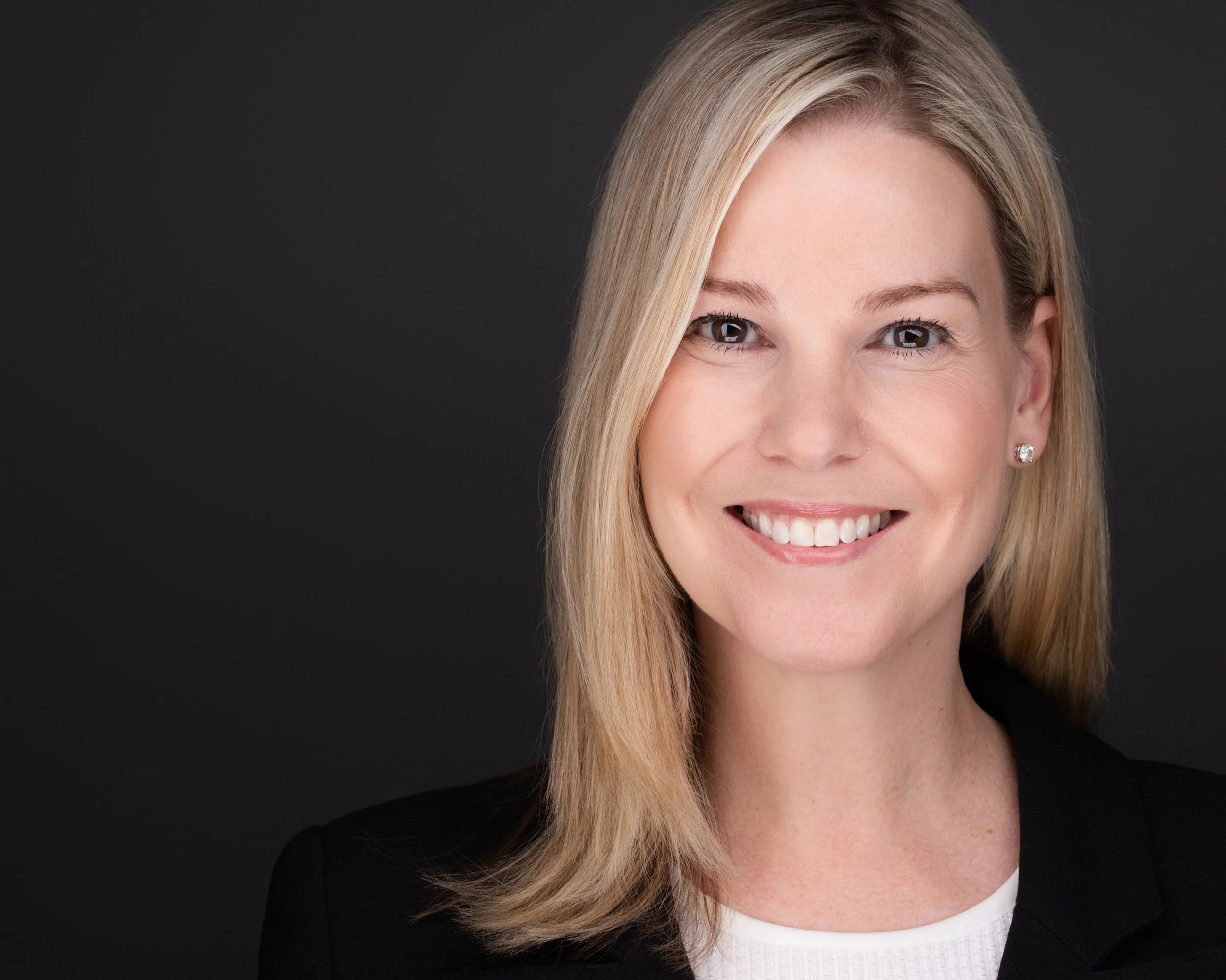 Professional woman with blonde hair, smiling, wearing a black blazer and pearl earrings, against a dark background.
