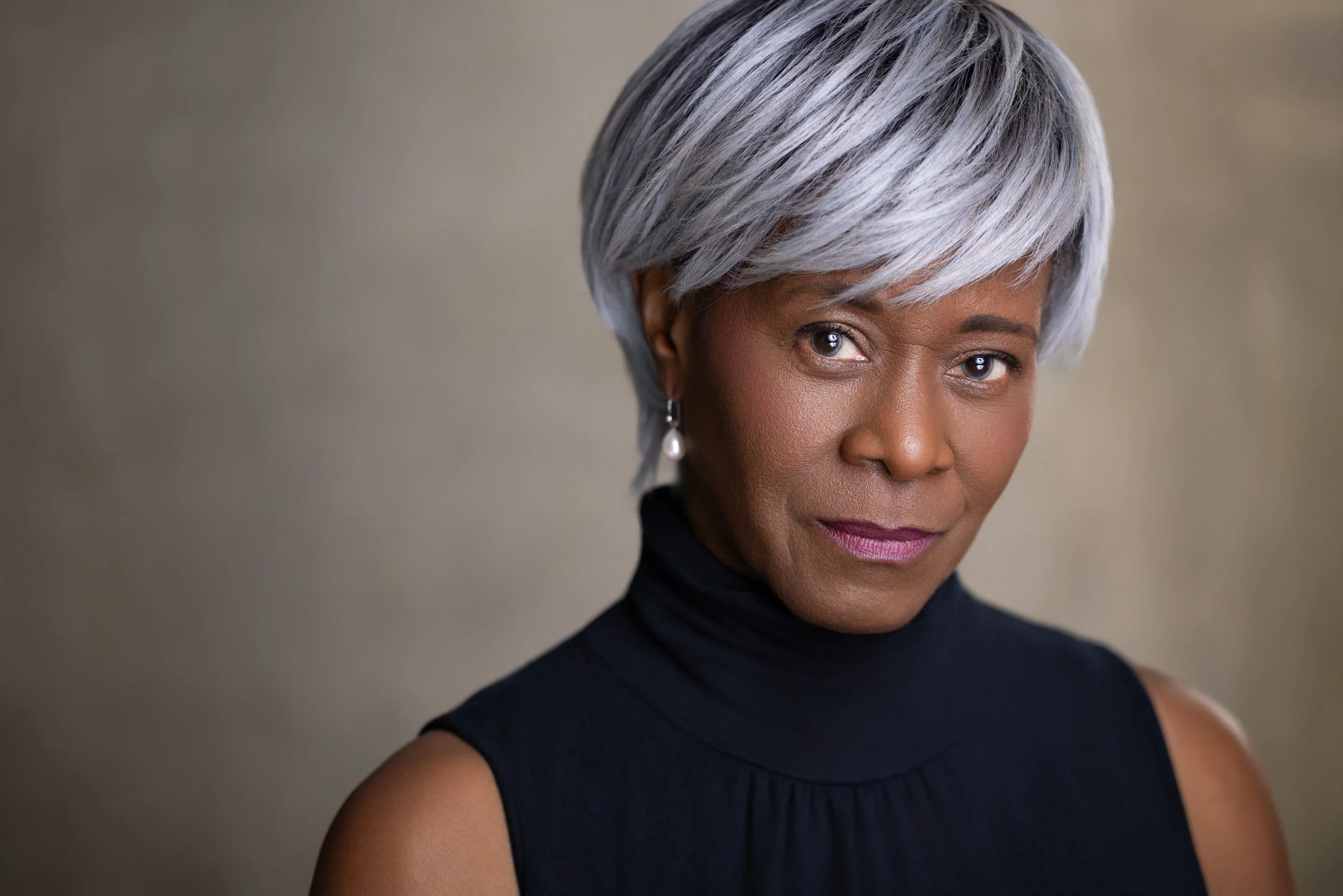 Portrait of an elegant woman with short silver hair, wearing earrings and a sleeveless black turtleneck, looking confidently at the camera.