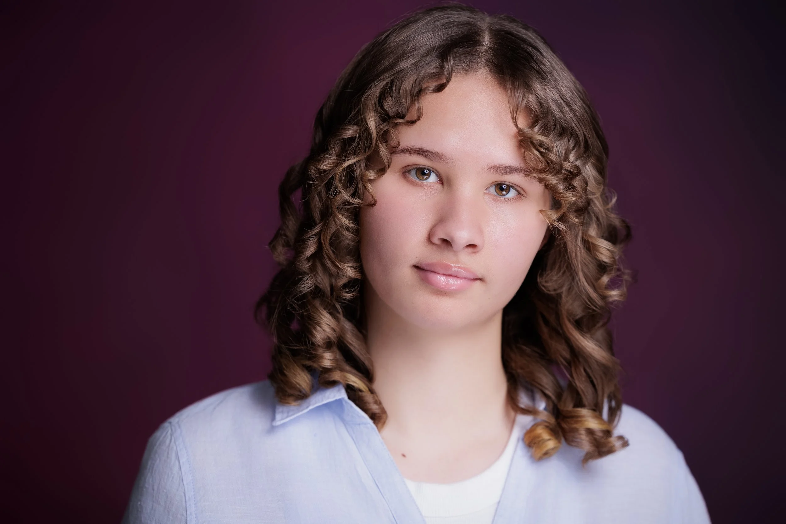 Portrait of a young woman with curly brown hair, wearing a light blue shirt, against a dark purple background.
