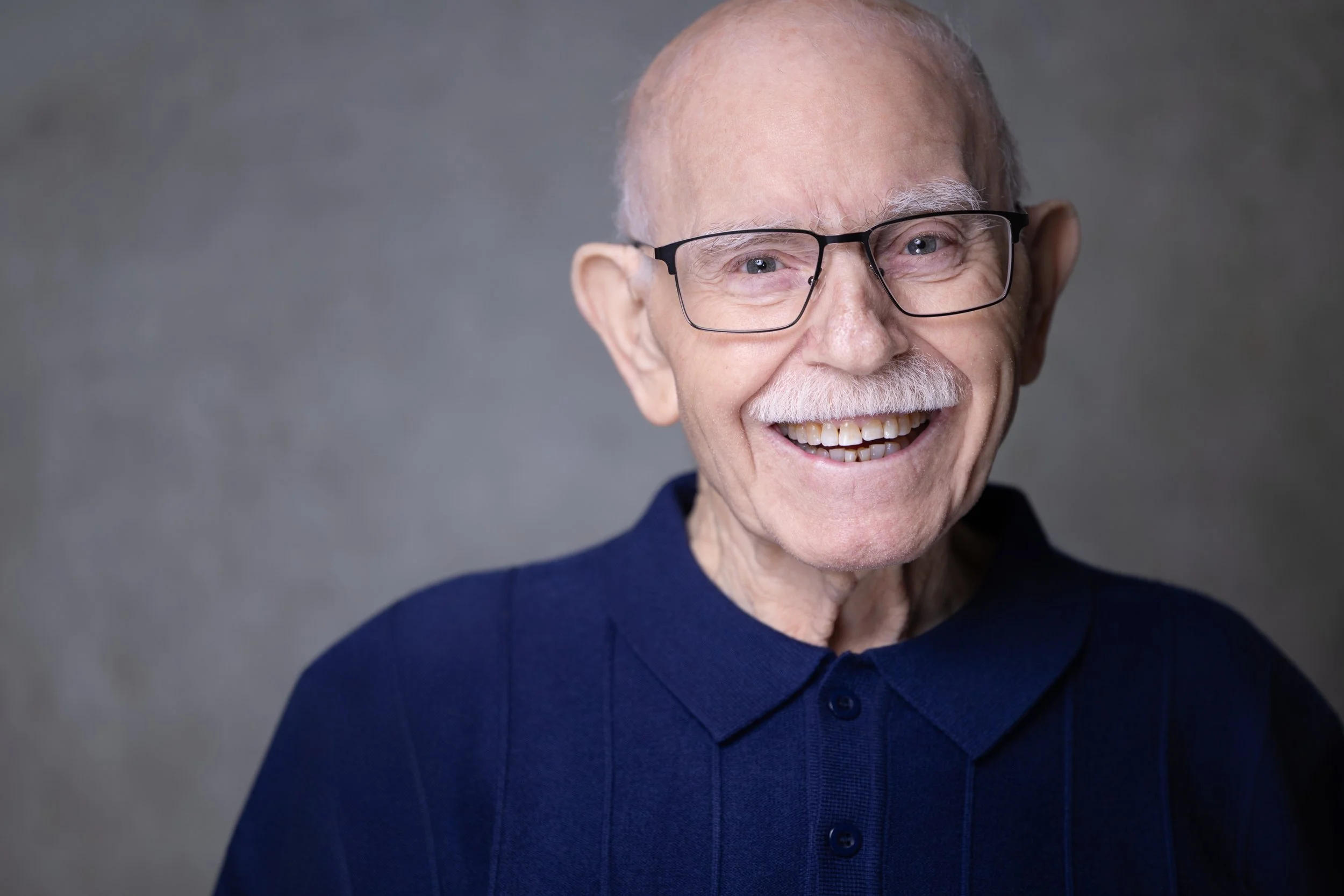 Smiling elderly man wearing glasses and a navy polo shirt, with a neutral gray background.