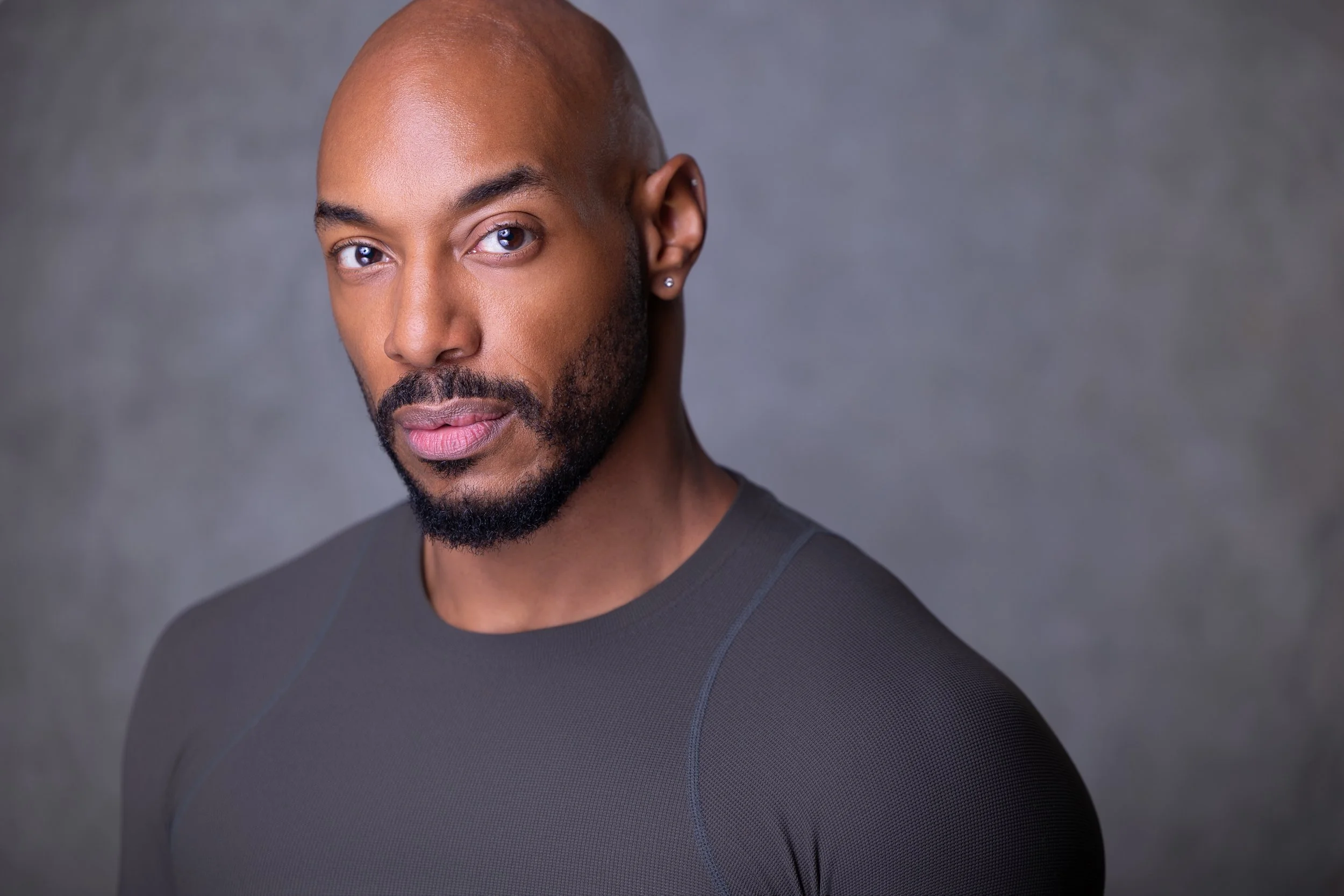 Close-up of a Black man with a shaved head and beard, wearing a dark athletic top, looking at the camera against a gray background.