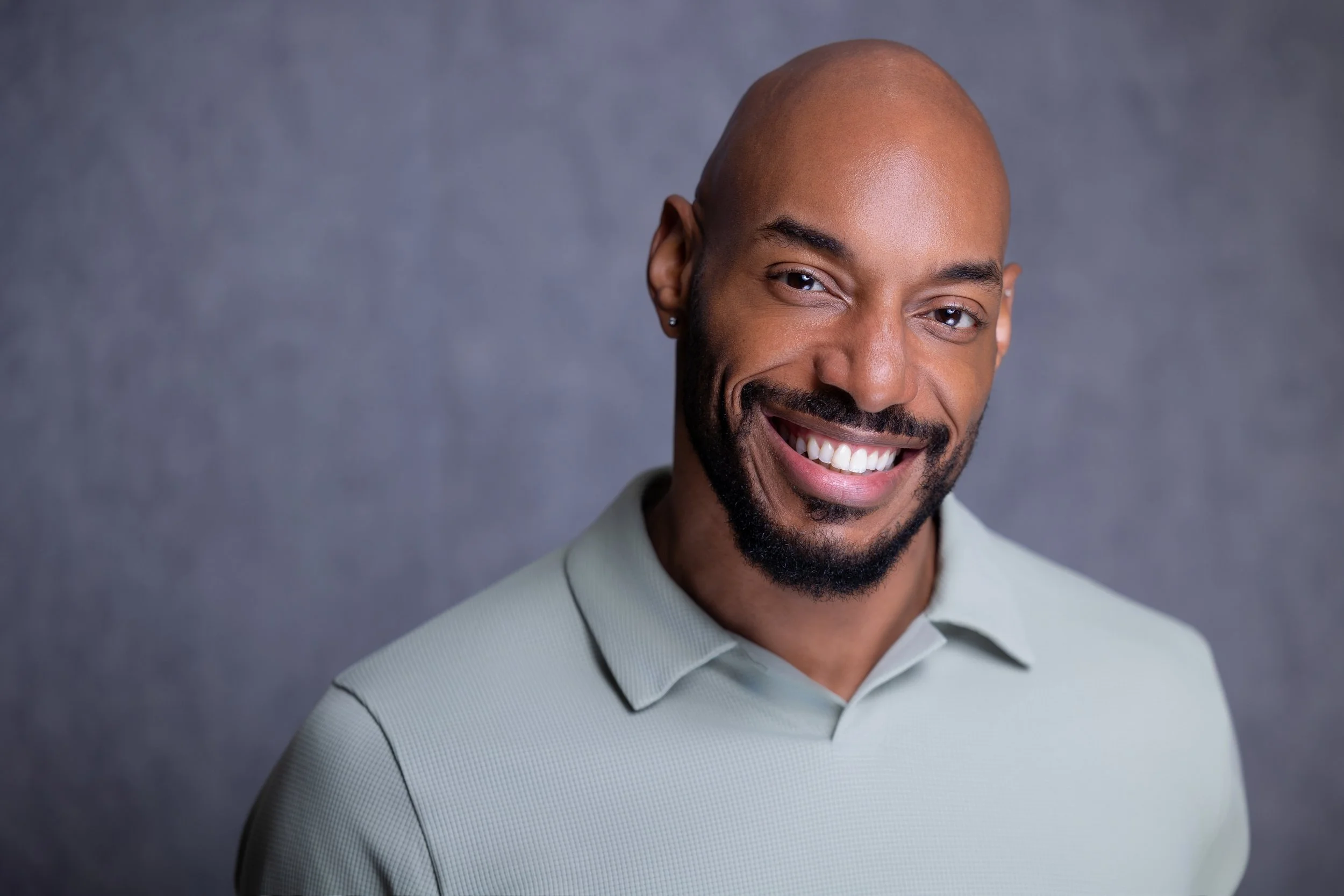 A smiling man with a bald head, beard, and mustache, wearing a light gray polo shirt, posing against a dark gray background.