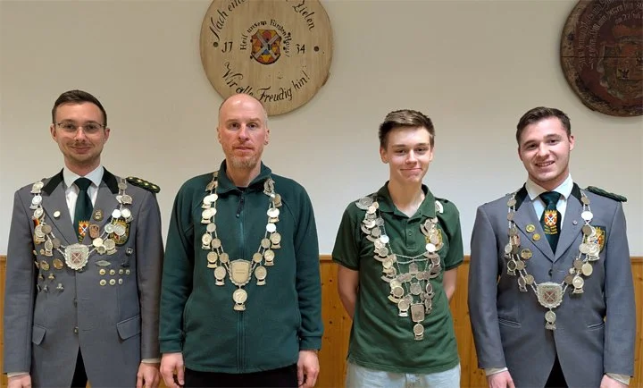 Vier junge Männer stehen nebeneinander, alle tragen Uniforme mit verschiedenen Orden und Ketten, in einem Raum mit Wanddekoration im Hintergrund.
