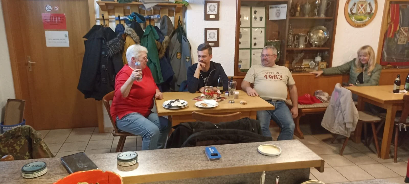 Vier Personen sitzen an einem Esstisch in einem gemütlichen Raum, während sie Getränke und Essen genießen, im Hintergrund eine geheime Wand mit Jacken und eine Glasvitrine.