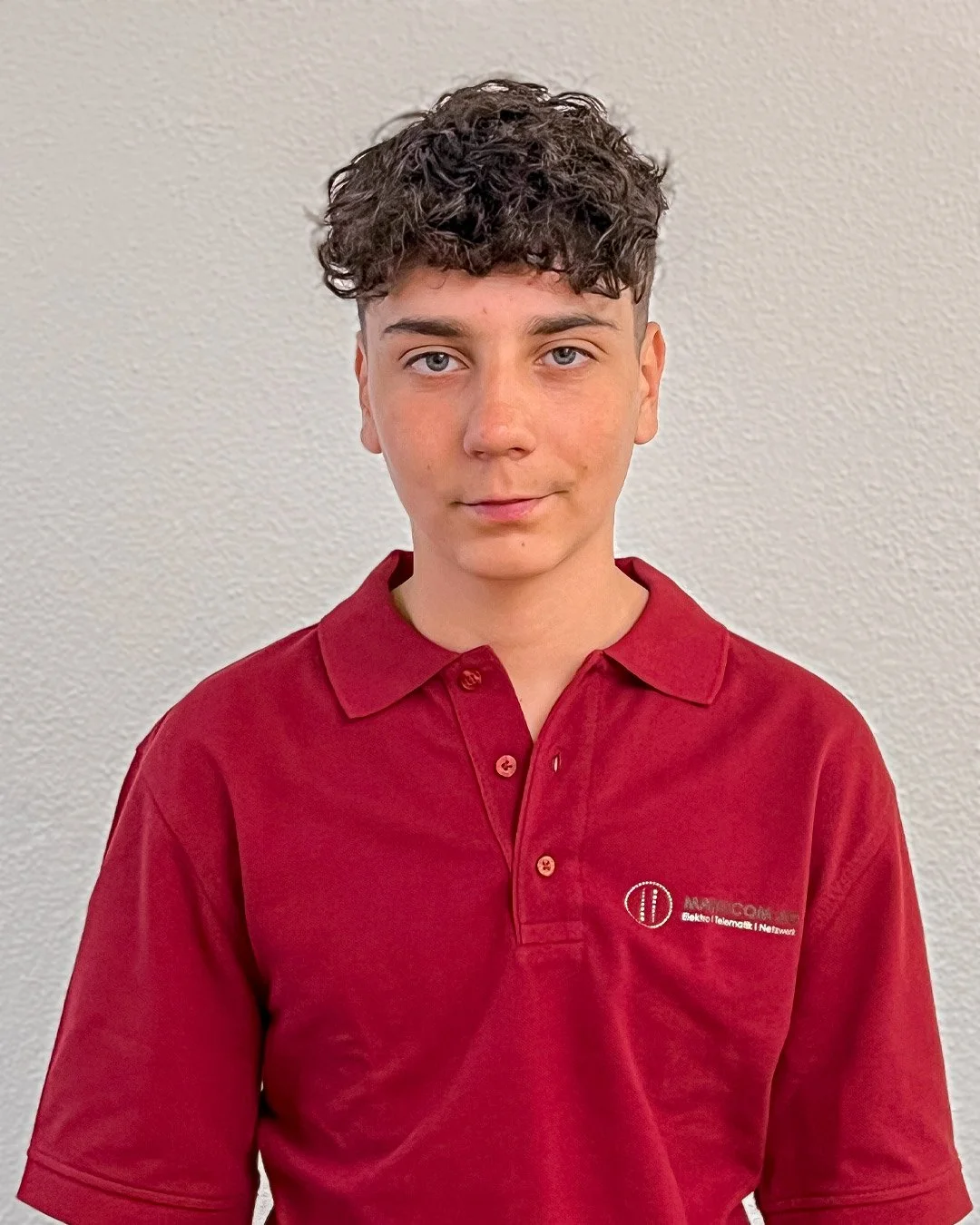 Junge trägt rote Poloshirt mit Logo, blickt in die Kamera, kurzer lockiger Brauner Haare, heller Teint, neutraler Gesichtsausdruck, geschlossene Lippen, gegen weiße Wand