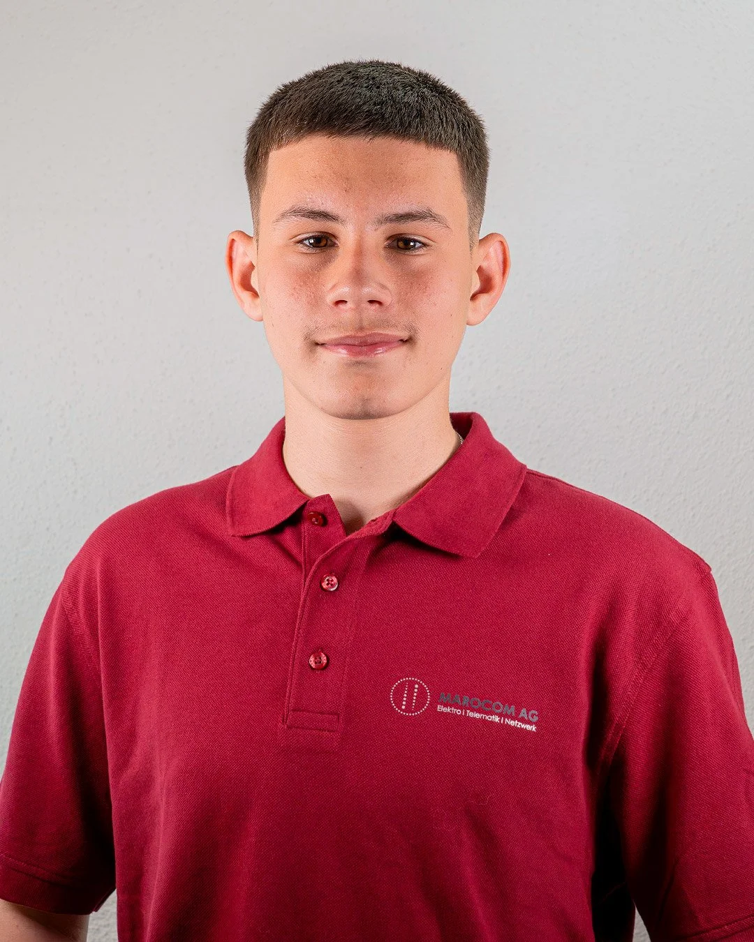 Junge in rotem Polo-Shirt mit gesticktem Firmenlogo steht vor grauer Wand.