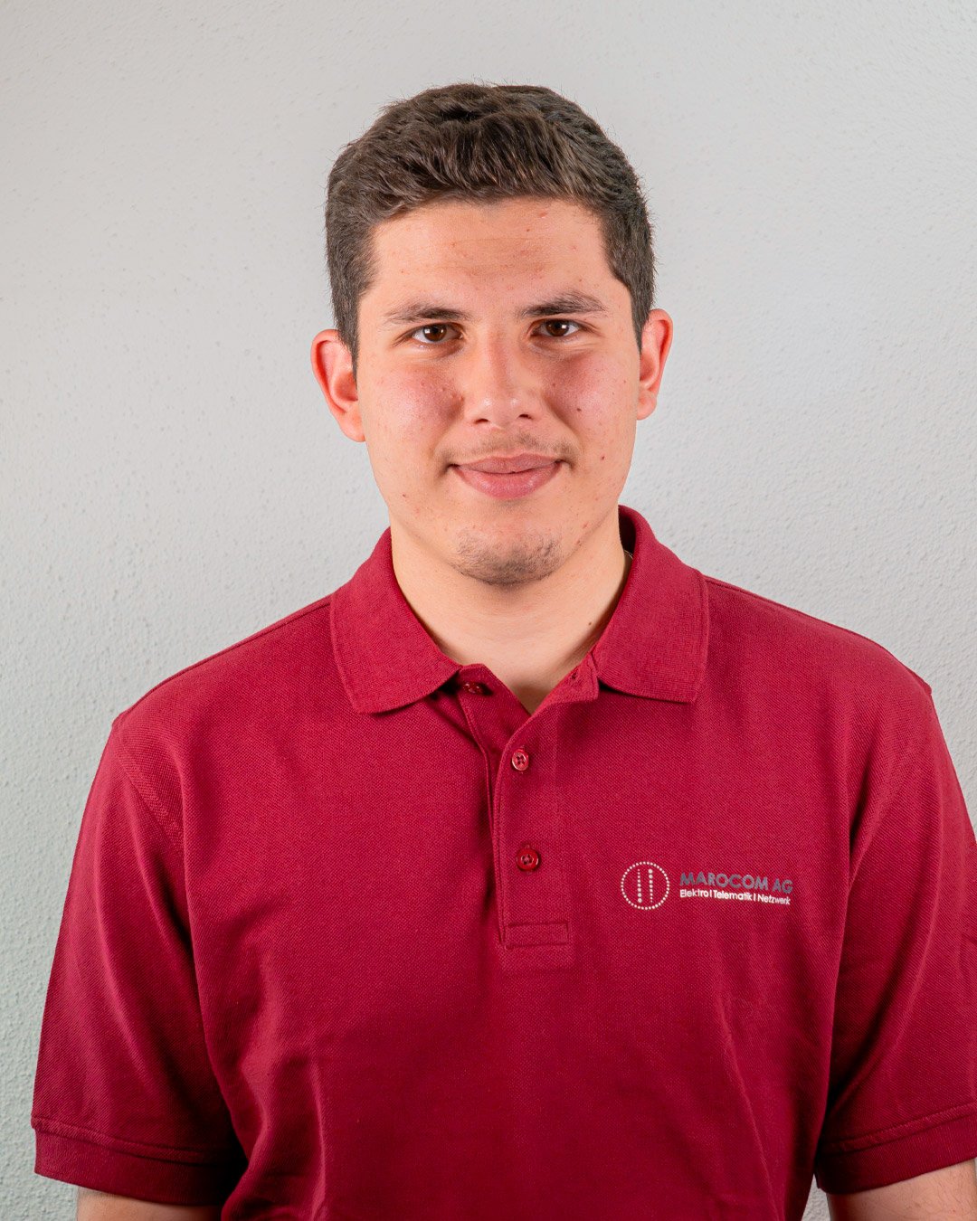 Junge trägt einen roten Polo mit Firmenlogo, steht vor grauer Wand.