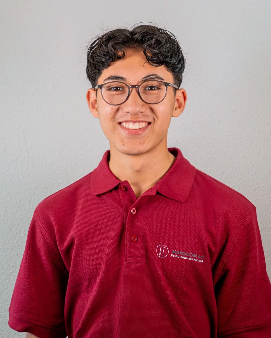 Junge mit curly Haaren und Brille, trägt ein rotes Polo-Shirt mit Firmenlogo, steht vor einer hellen Wand, lächelt in die Kamera.