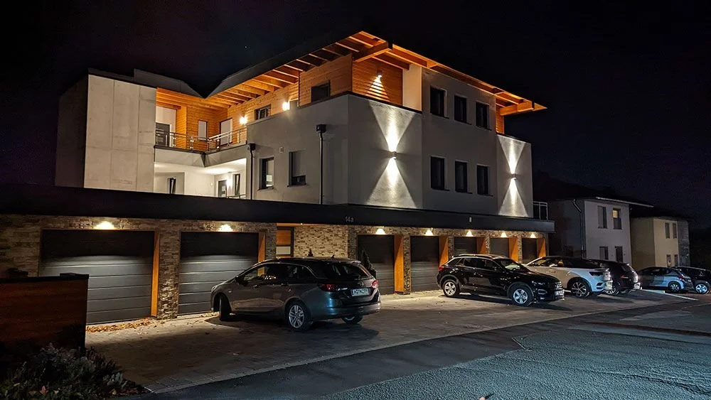 Modernes Mehrfamilienhaus bei Nacht mit beleuchteten Außenwänden und mehreren geparkten Autos in der Einfahrt.