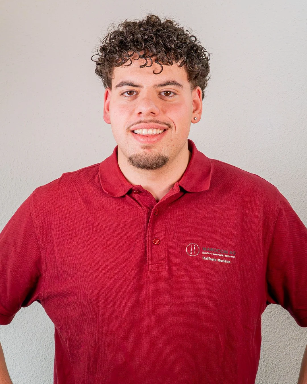 Junger Mann mit dunklen, lockigen Haaren, Vollbärtchen und einem Ohrpiercing, trägt ein rotes Polo-Shirt mit Firmenlogo und Name, steht vor heller Wand.