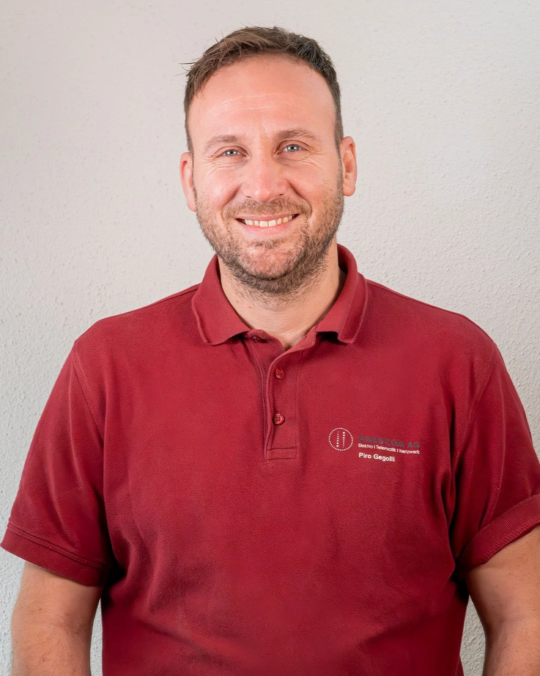 Ein lächelnder Mann mit kurzem braunem Haar, Bart und blauen Augen trägt ein rotes Polo-Shirt mit Firmenlogo auf der linken Brustseite, das den Namen Piro Gecolli enthält, vor einer cremefarbenen Wand.