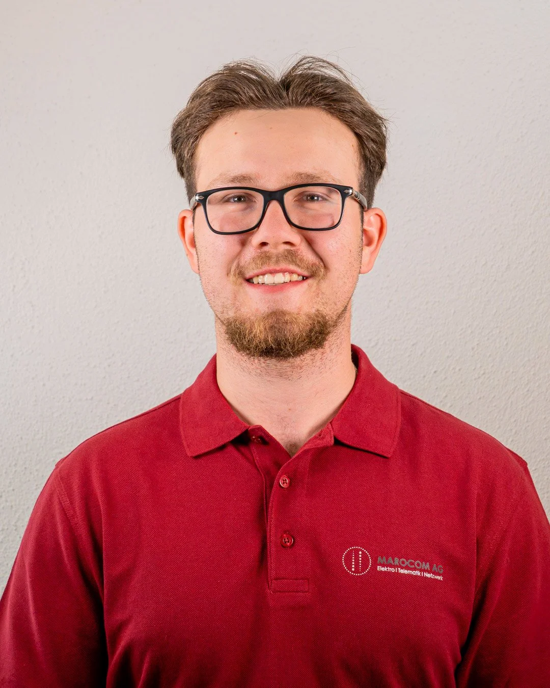 Mann trägt rotes Polohemd mit Firmenlogo, lächelt, gegen weißen Hintergrund.