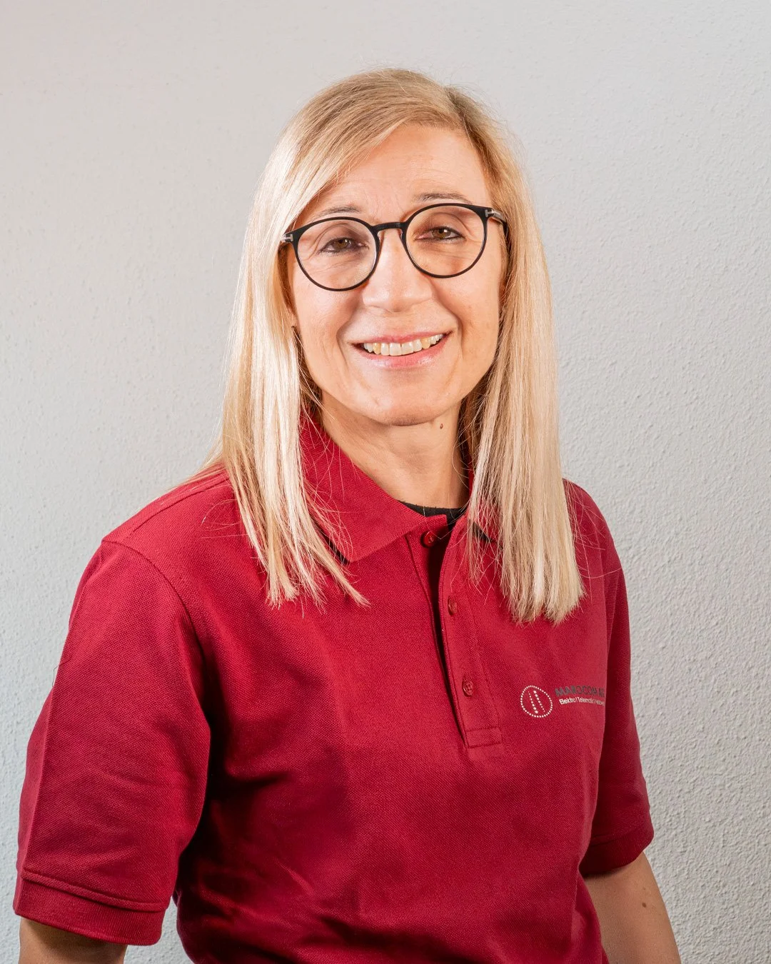 Frau mit Brille und roten Polo-Shirt, lächelnd vor weißer Wand.