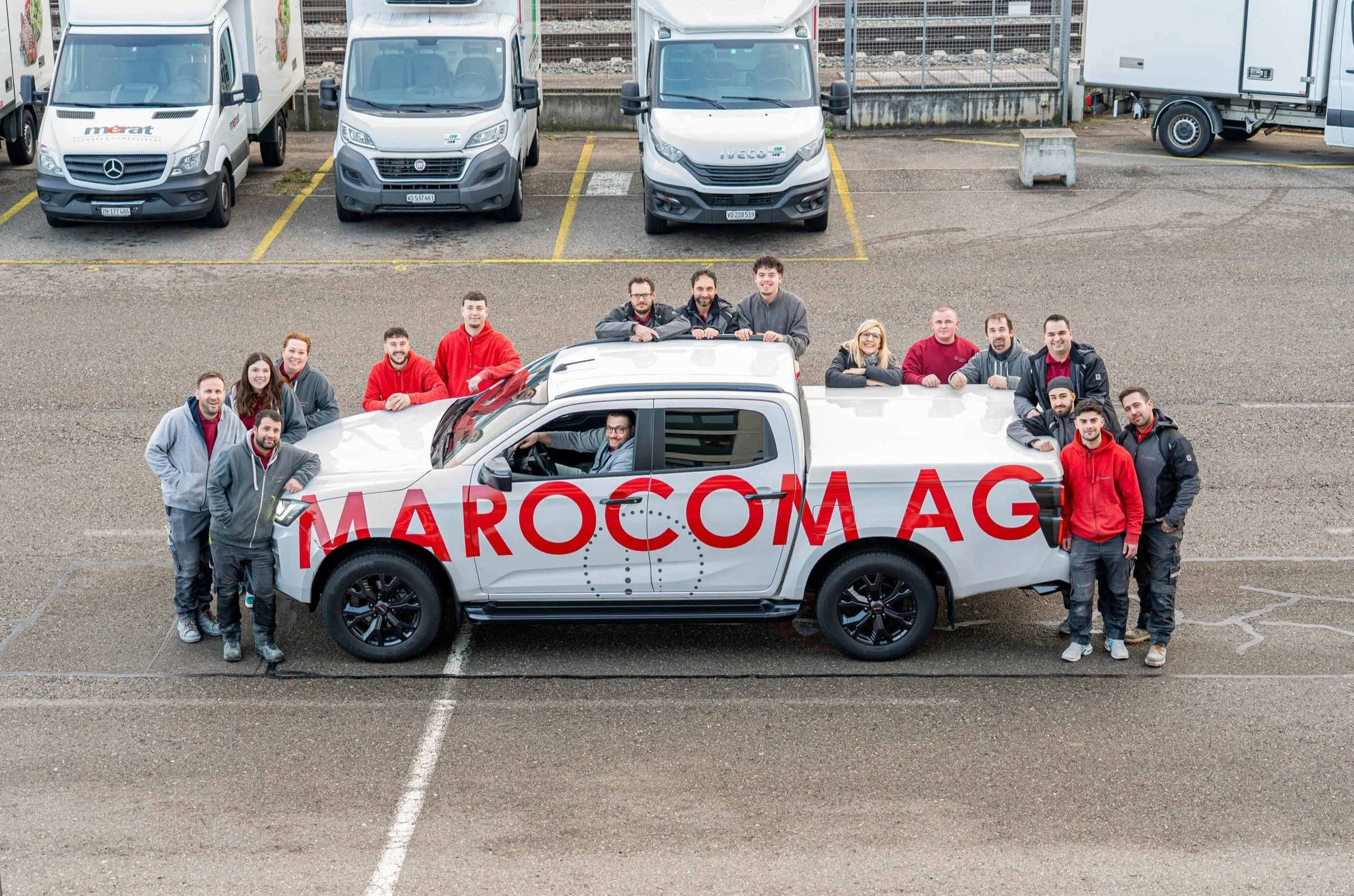 Gruppenfoto von Menschen rund um einen weißen Lieferwagen mit roter Schrift 'MAROCOM AG' in einem Parkplatz, mit mehreren parkenden Fahrzeugen im Hintergrund.