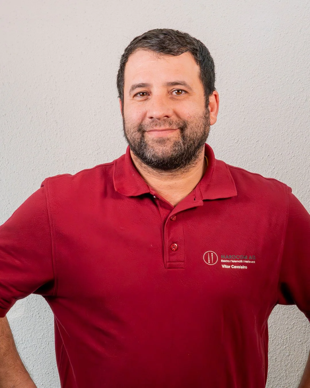 Ein Mann trägt ein rotes Polo-Shirt mit Logo und Text auf der linken Brustseite, steht vor einer hellen Wand, lächelt leicht, hat dunkles Haar und einen Bart.