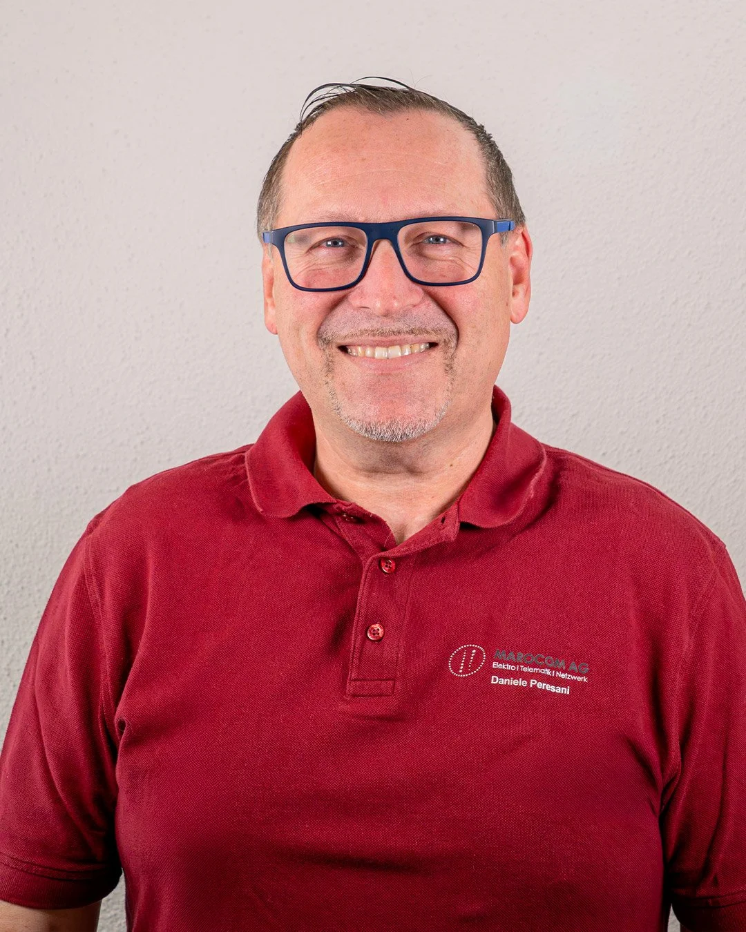 Ein Mann trägt eine rote Polo-Shirt mit Logo und Namen, trägt blaue Brille und hat ein Lächeln, steht vor einer weißen Wand.