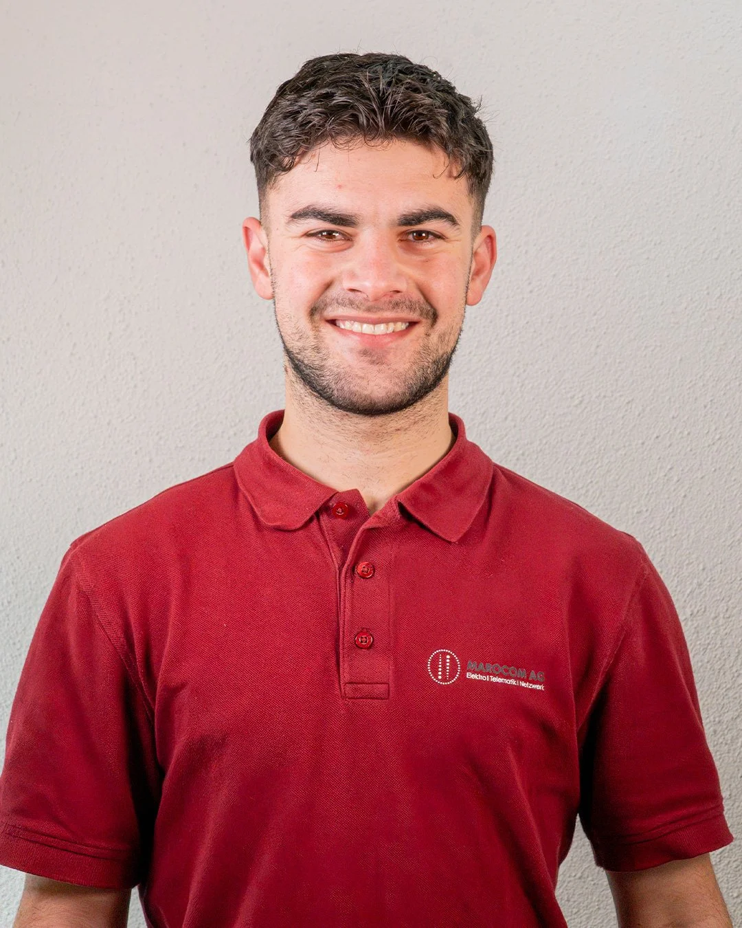 Junge Mann trägt rote Polo-Shirt mit Firmenlogo, lächelt vor heller Wand.
