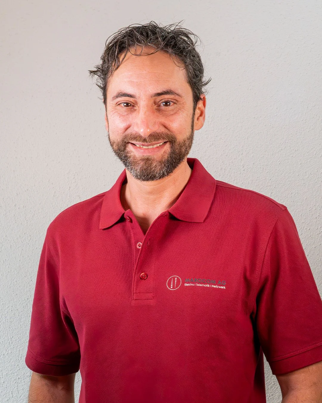 Ein Mann mit braunem Haar, Bart und freundlichem Lächeln trägt ein rotes Polo-Shirt mit Logo und Schriftzug auf der linken Brust, steht vor einer neutralen hellgrauen Wand.