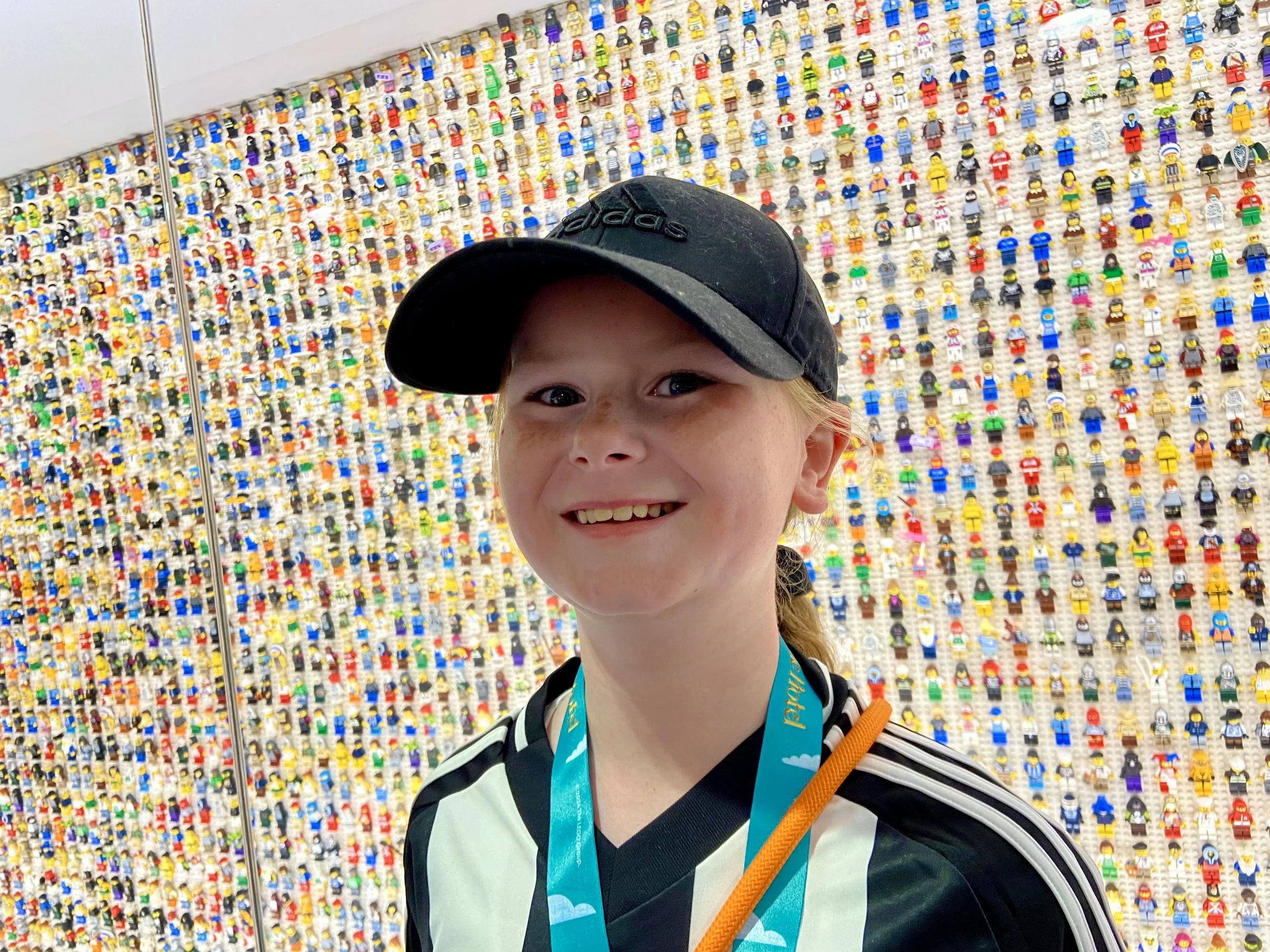 A smiling young girl with a black Adidas cap and a black and white sports jersey standing in front of a wall filled with colorful LEGO minifigures.