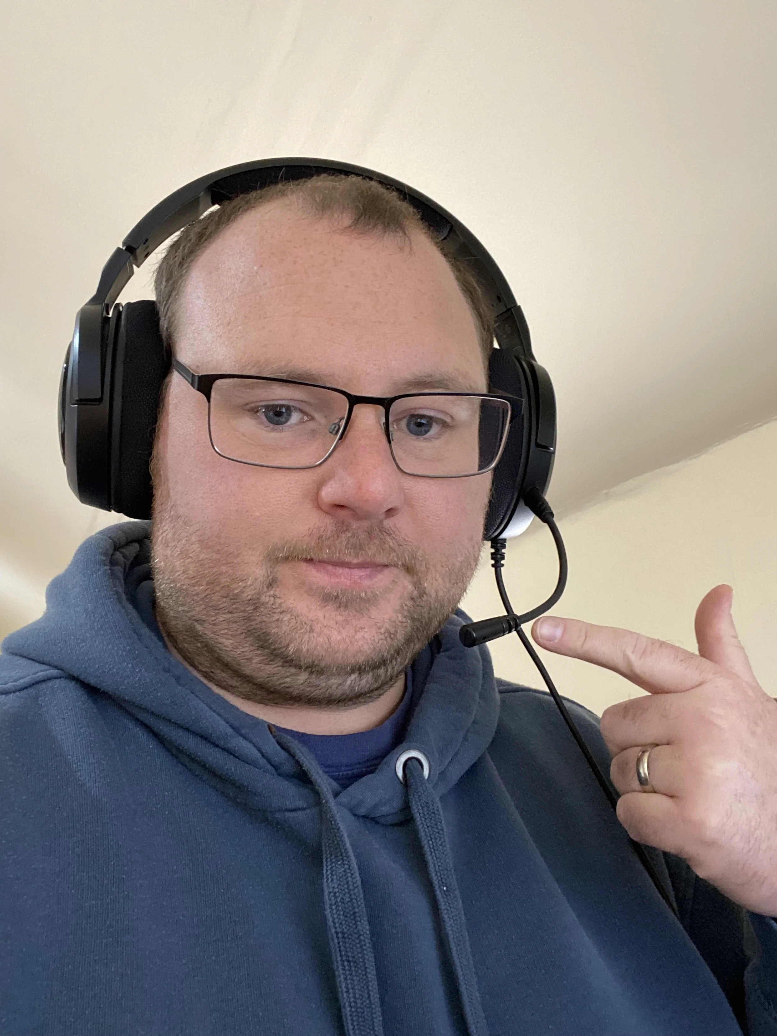 A man with glasses, a beard, and short hair wearing a dark blue hoodie and large black headphones, pointing at the headphones with his index finger.