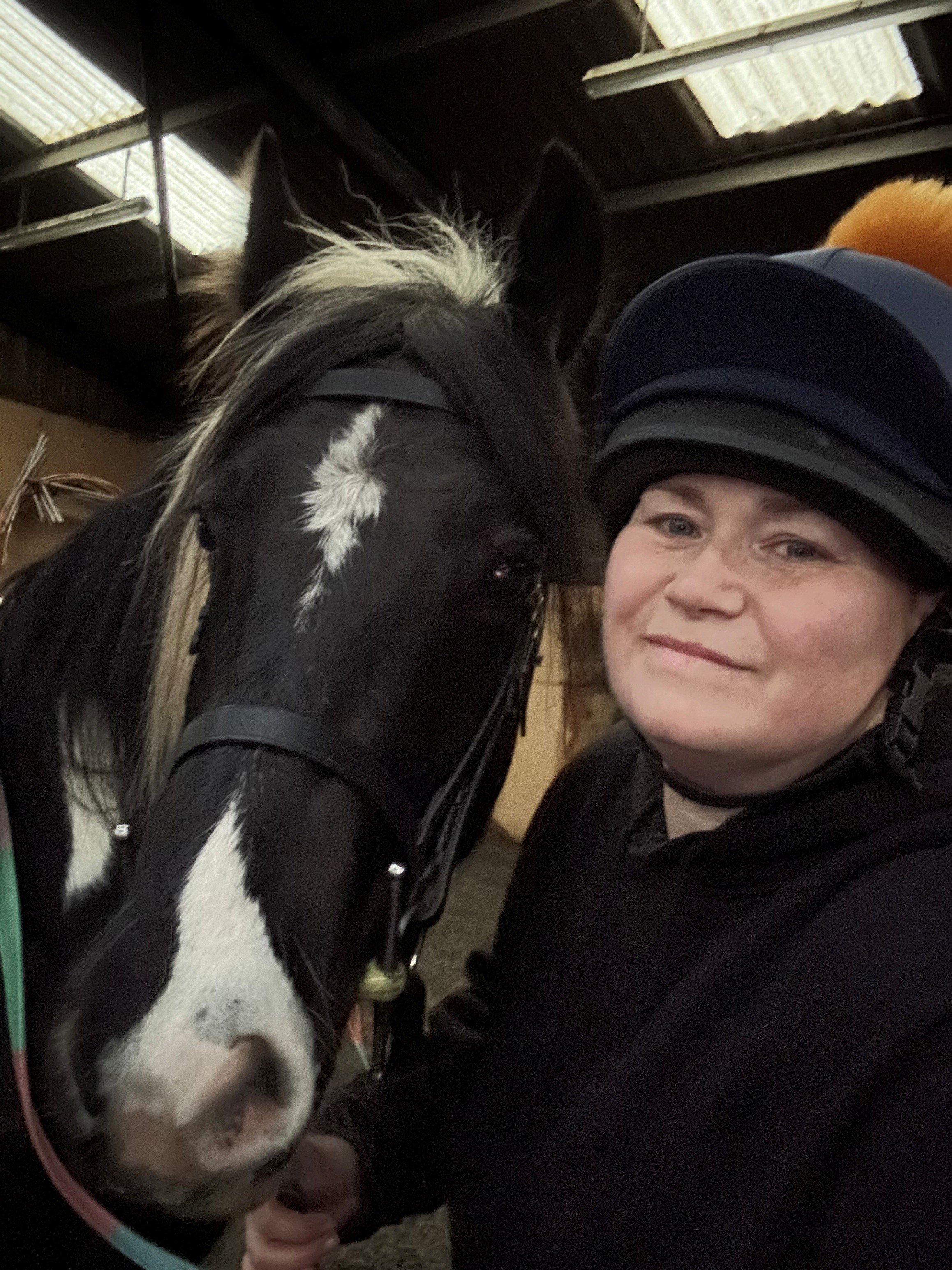 A woman in riding gear and a helmet taking a selfie with a black and white horse inside a stable.