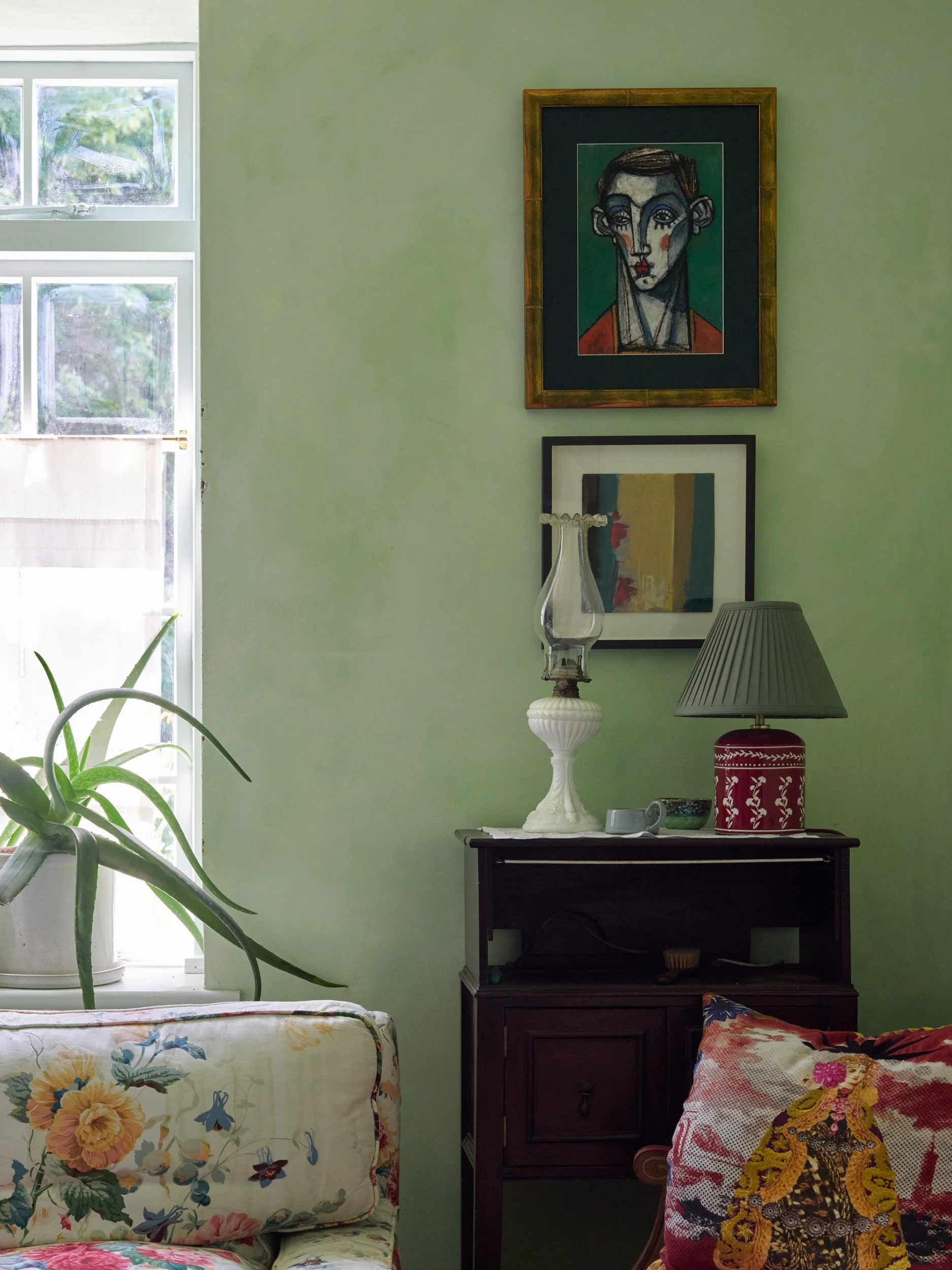 An interior living room corner with a light green wall, a window with white frames, a sofa with floral upholstery, a dark wooden cabinet with a vintage oil lamp, a red and white patterned table lamp, framed artwork, and a potted plant.