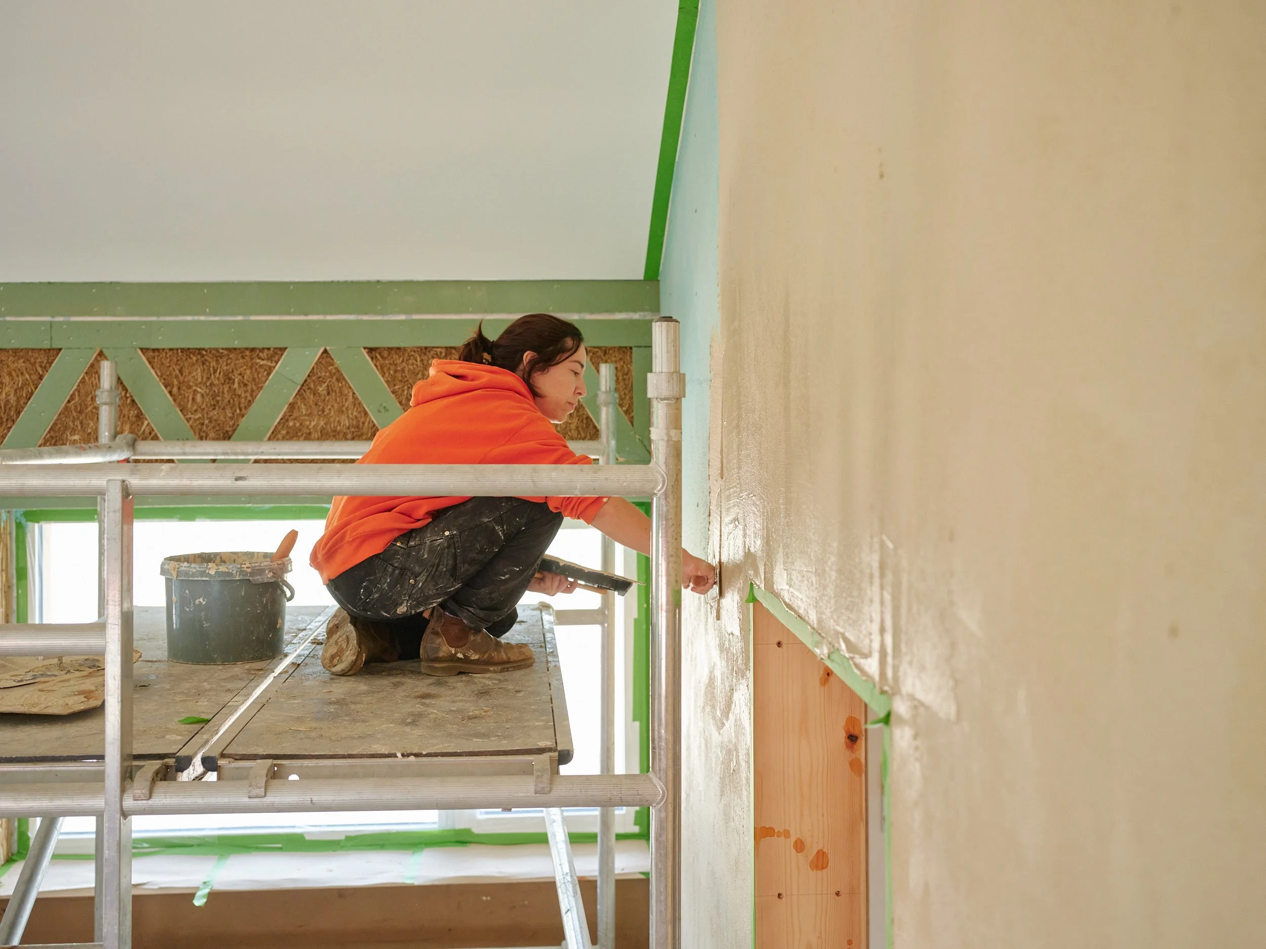 A woman in an orange hoodie is squatting on a scaffolding while painting a wall in a room under construction. She is using a paint roller and has a bucket of paint nearby.