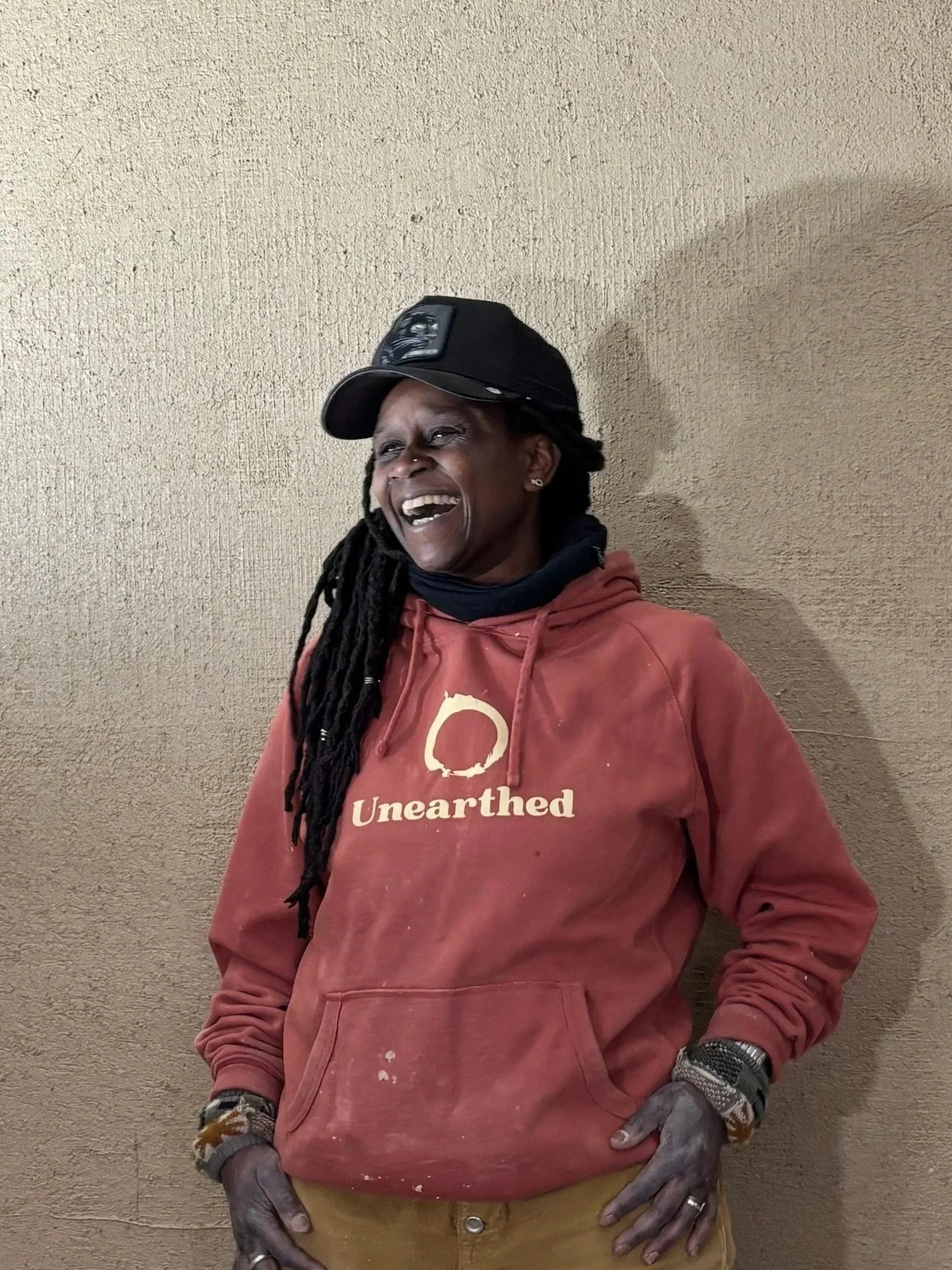 A person with long dreadlocks wearing a gray cap, black t-shirt, and khaki pants, smiling and sitting on a log outdoors with green grass and trees in the background.