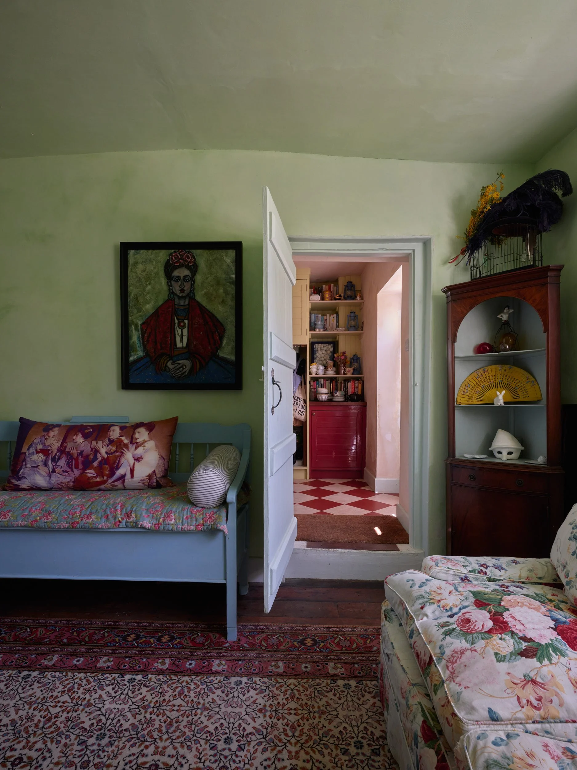 A cozy living room with green walls, a floral sofa, a turquoise bench with a decorative pillow, and a wooden cabinet with decorative items. An open door reveals a kitchen with red and white checkered flooring and shelves with jars and books.