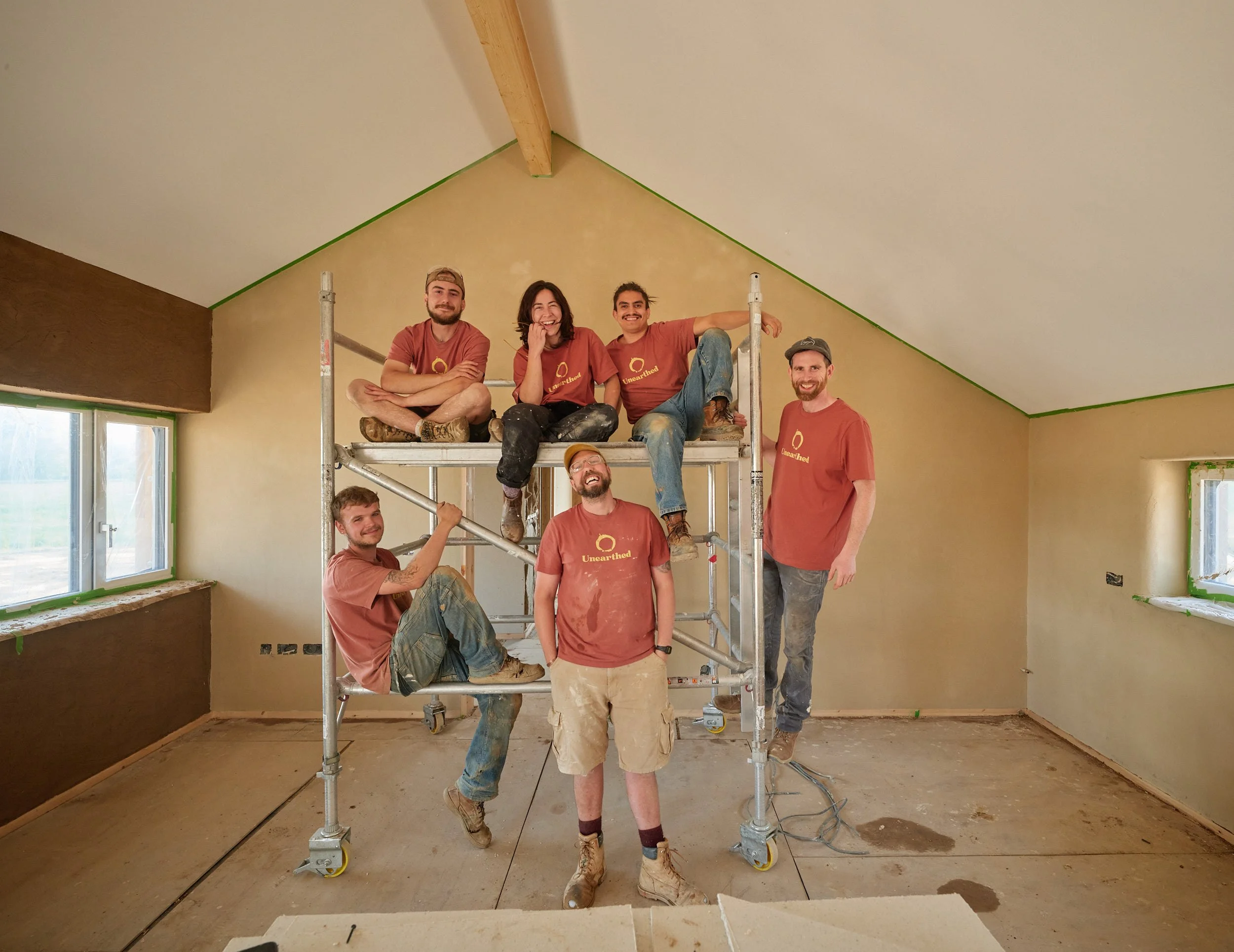 Group of eight people, all wearing matching t-shirts, posing on and around scaffolding inside a room under construction or renovation. Some are sitting on the scaffolding, while others stand beside it, smiling and relaxed.