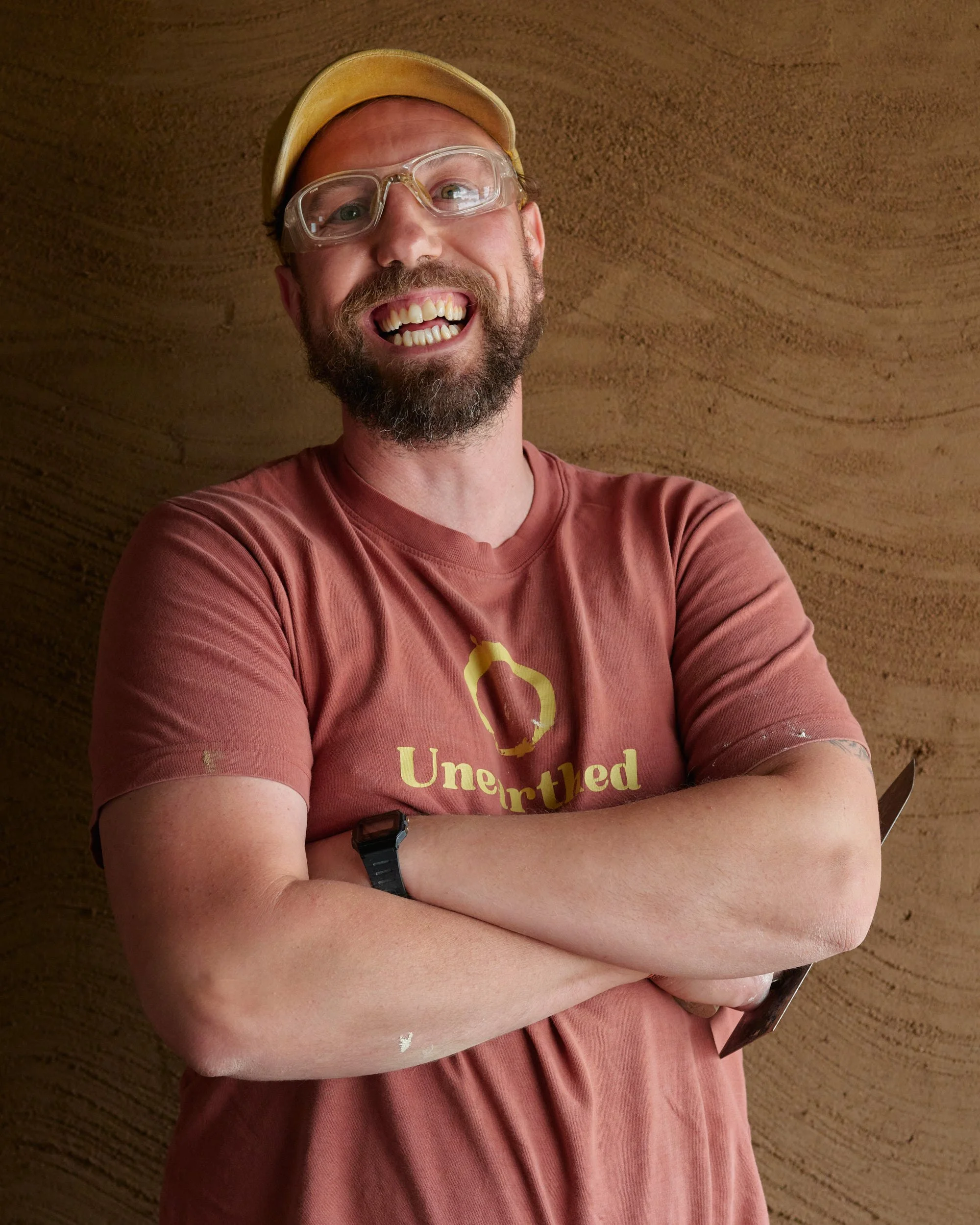 A smiling man with a beard wearing safety goggles, a beige cap, and a brown t-shirt with white paint splatters.