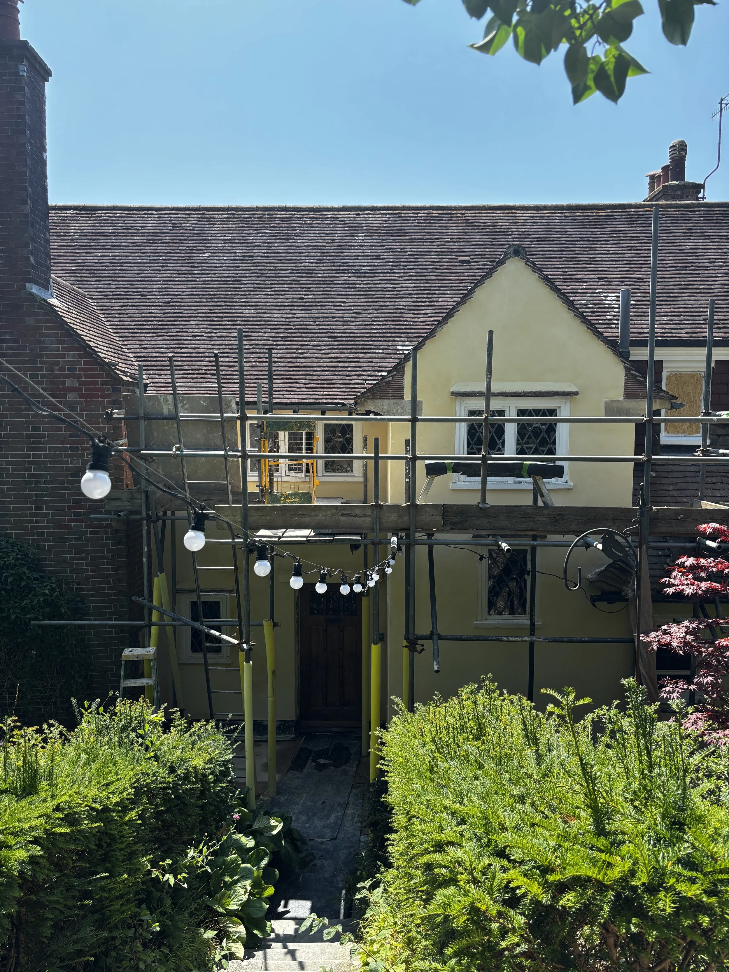 A house showing completed exterior renovation with scaffolding, string lights, and a garden in the foreground.