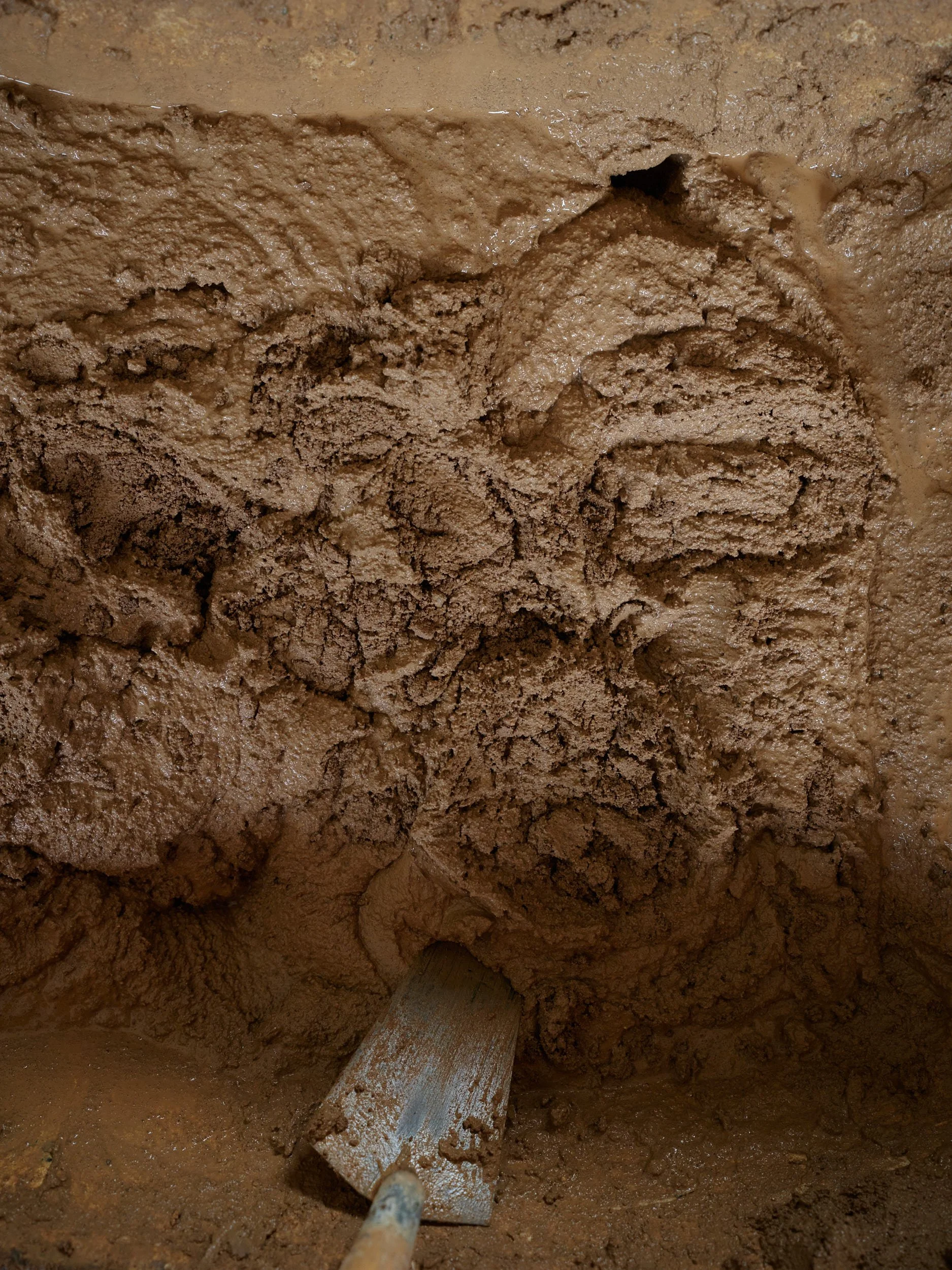 Close-up of wet, brown clay or mud being mixed or shaped with a flat wooden tool.