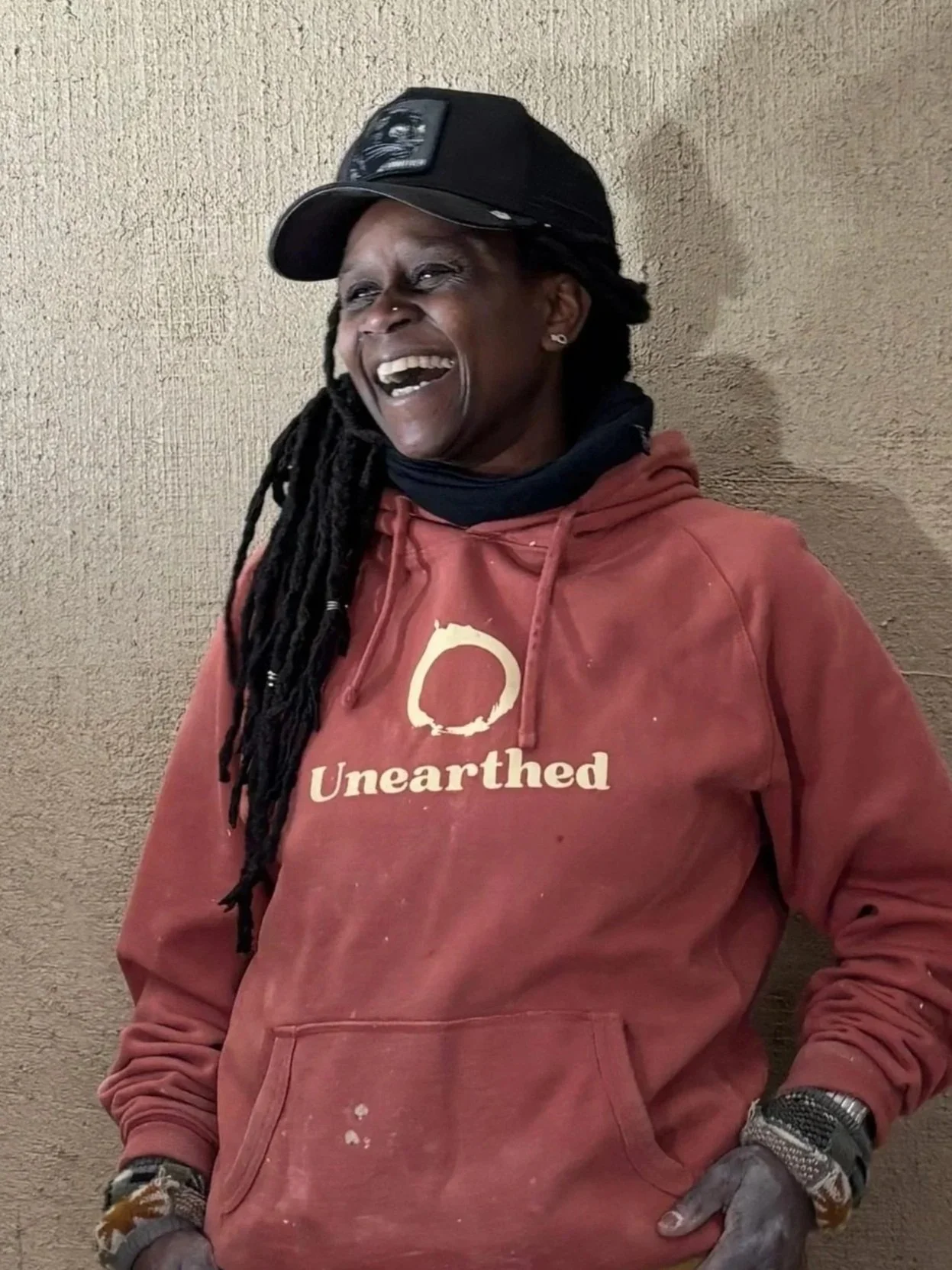 A person with long dreadlocks wearing a gray cap, black t-shirt, and khaki pants, smiling and sitting on a log outdoors with green grass and trees in the background.